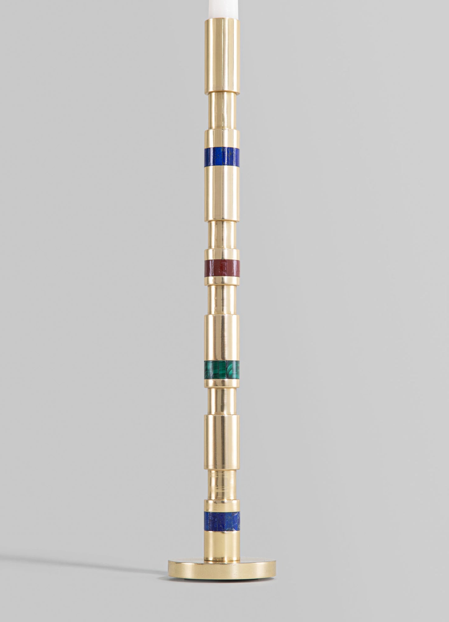 A tall brass candlestick with a cylindrical base and a layered, segmented design featuring colored rings in blue, red, and green, with a white candle on top. The style appears modern with geometric elements.