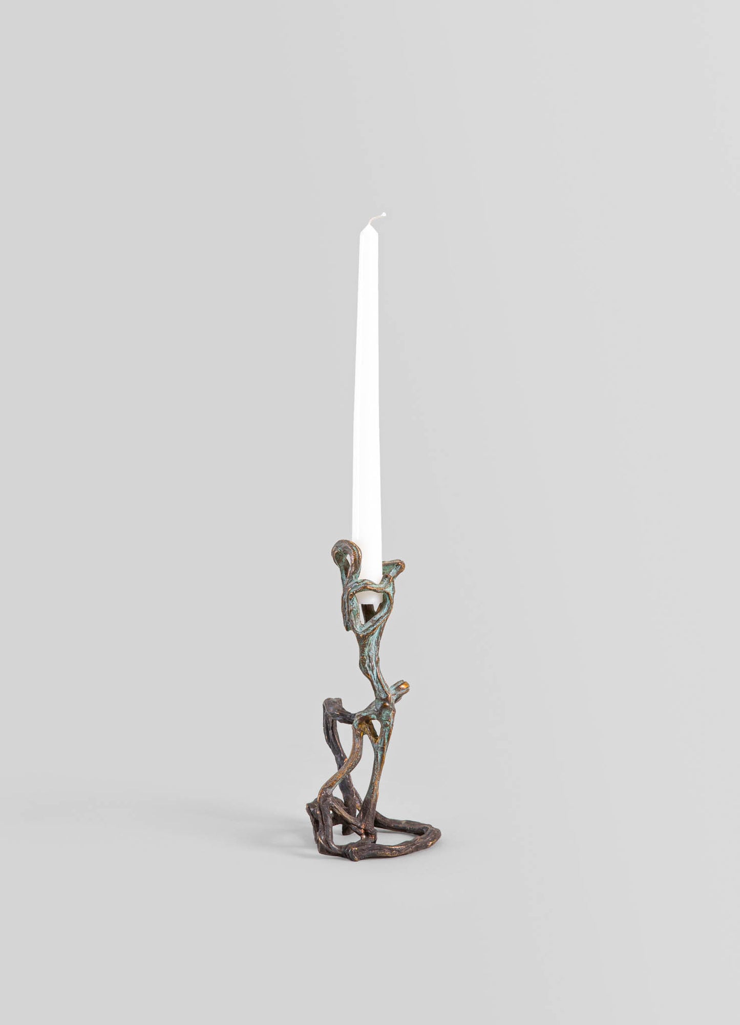 A decorative candle holder made of metal with a bronze finish, featuring an organic, sculptural design resembling intertwined branches or roots. It holds a tall, white taper candle.