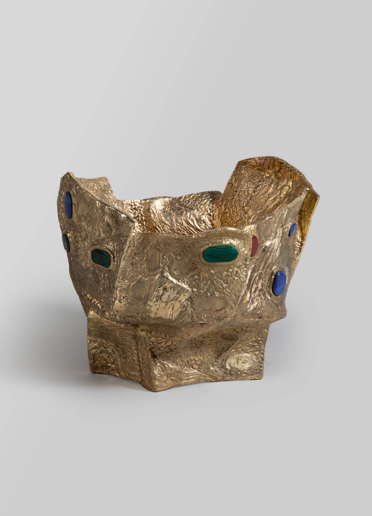 A decorative bowl made of textured gold-colored metal with an irregular, sculptural shape. It features oval-shaped inlays in blue, green, and red, creating a bold, artistic design.