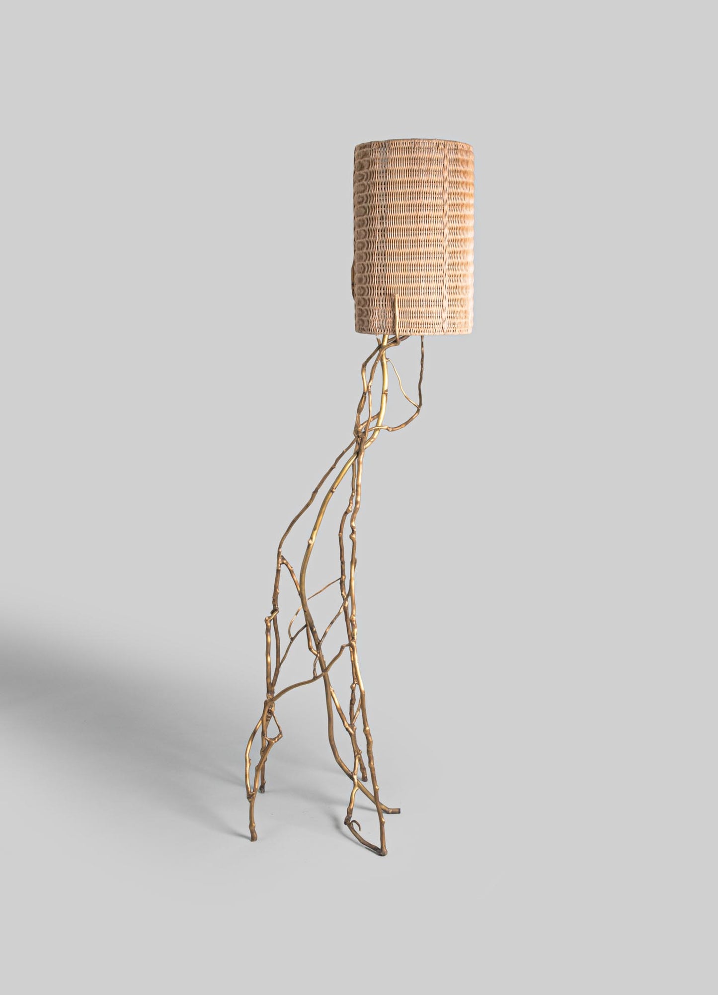 A floor lamp with a brass, branch-like base and a drum-shaped shade made of woven rattan, featuring a natural, textured surface in neutral tones.