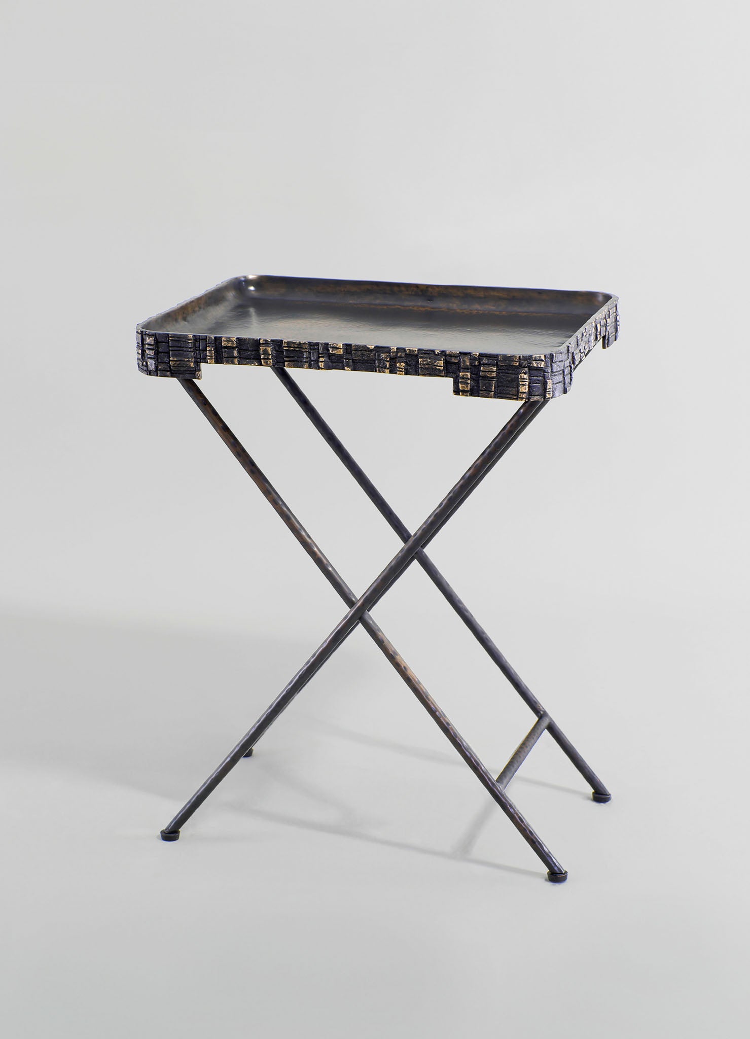 A small metal tray table with a rectangular black surface featuring a slightly textured finish, supported by crossed metal legs with a distressed, dark finish. The table has a rustic and industrial style.