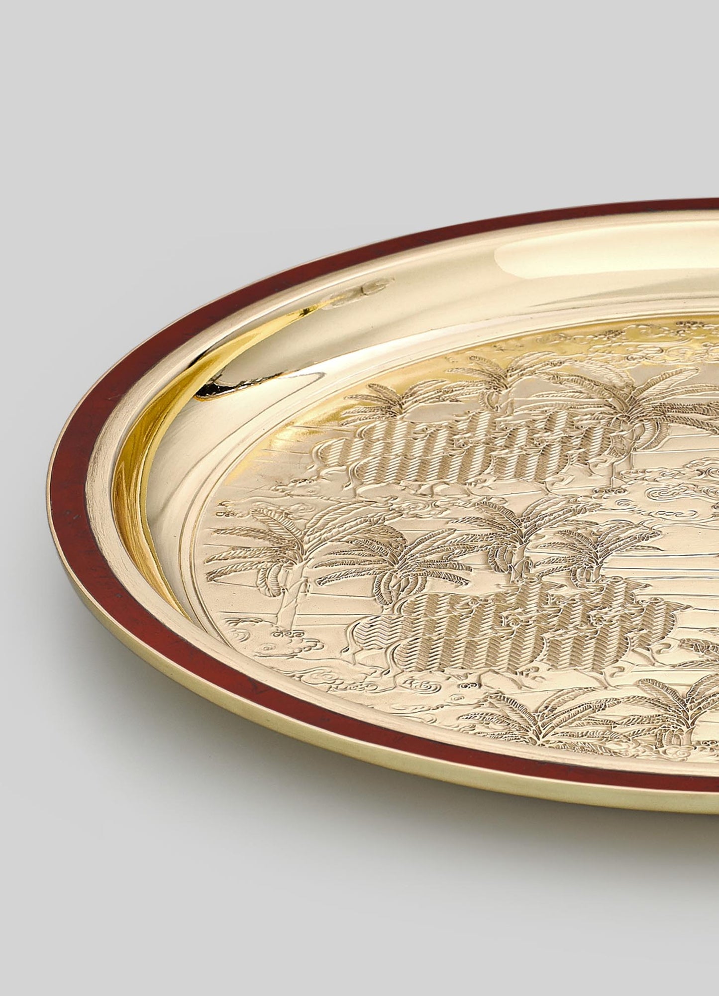 A round tray made of shiny gold metal with engraved palm trees and decorative motifs, edged with a dark red border. The design features detailed tropical scenery and intricate patterns, exuding a luxurious and vintage style.