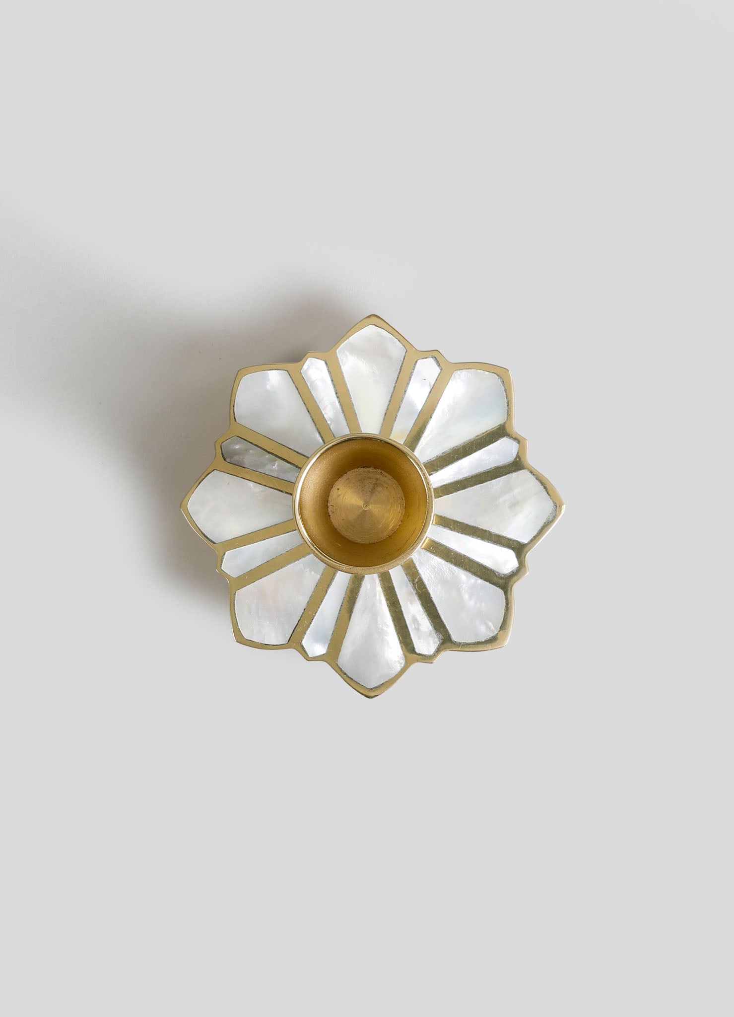 A decorative mirror with a flower-shaped design featuring multiple petal-like sections made of mother-of-pearl, framed in gold. The mirror is paired with a small brass holder or candle cup in the center, set against a light background.