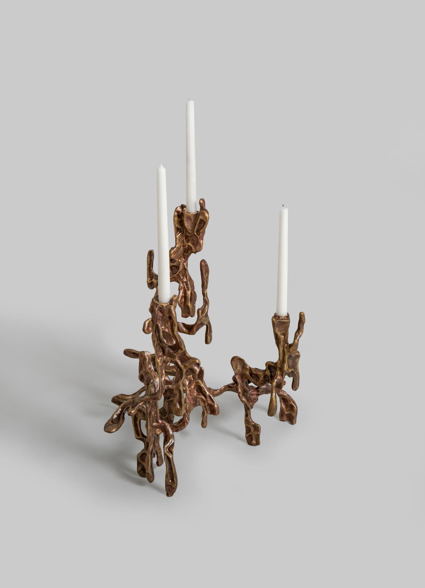 A sculptural candleholder made of metallic bronze material with an organic, abstract, and irregular design, holding three white taper candles. The piece has a contemporary style with a textured, freeform motif.