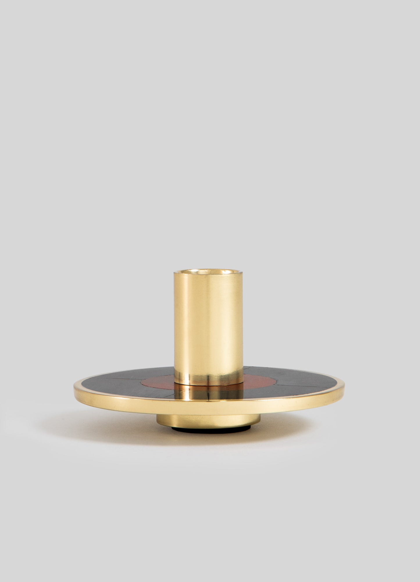 A modern brass candle holder featuring a cylindrical holder with a smooth finish, mounted on a round, flat base with black and copper accents for contrasting tones.