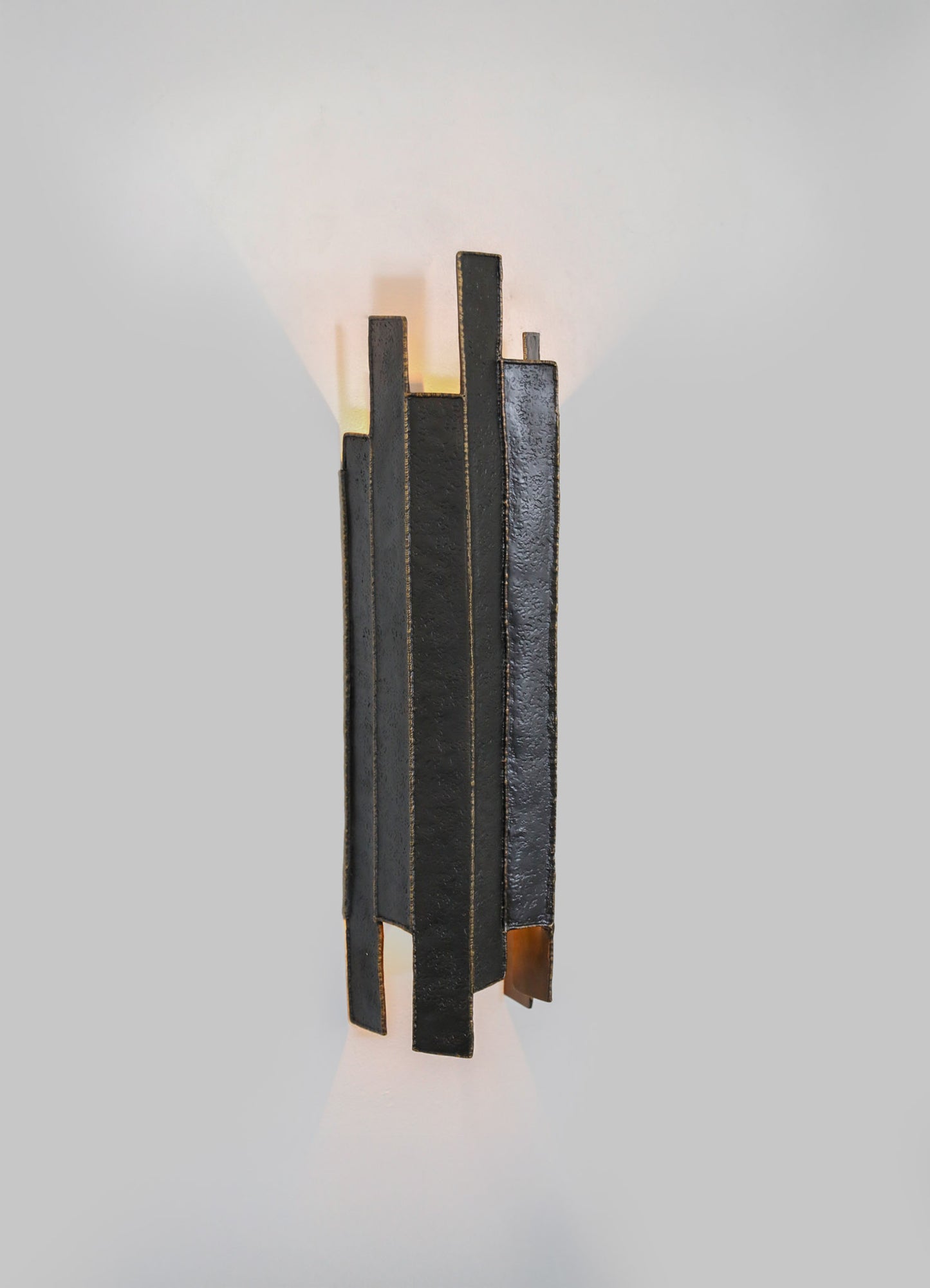 A modern black wall sconce with a textured metal finish, featuring geometric vertical panels and warm ambient lighting casting triangular shadows on a plain wall.