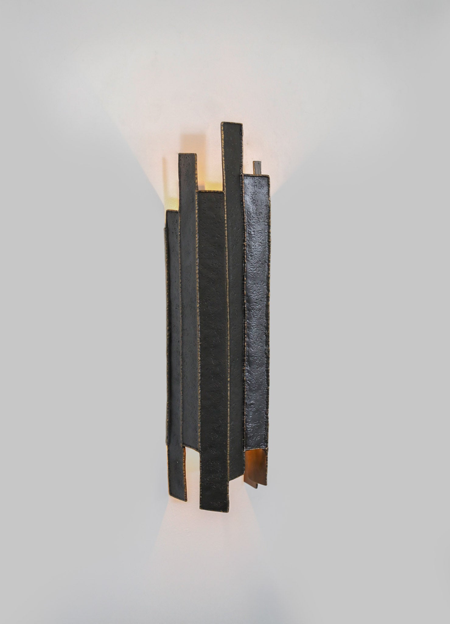 A modern black wall sconce with a textured metal finish, featuring geometric vertical panels and warm ambient lighting casting triangular shadows on a plain wall.