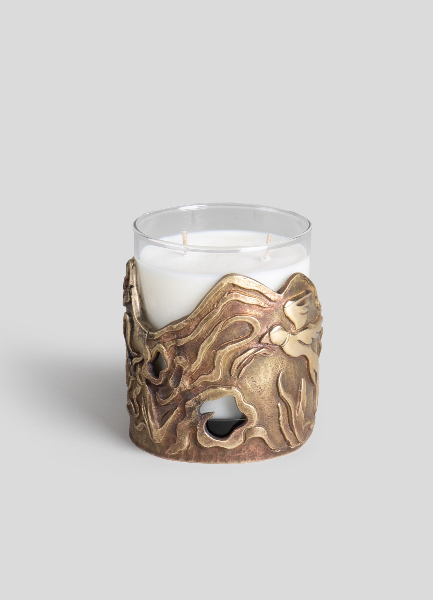 A glass candle holder with a decorative brass accent wrap featuring abstract, flowing lines and organic shapes, designed to hold a white pillar candle.