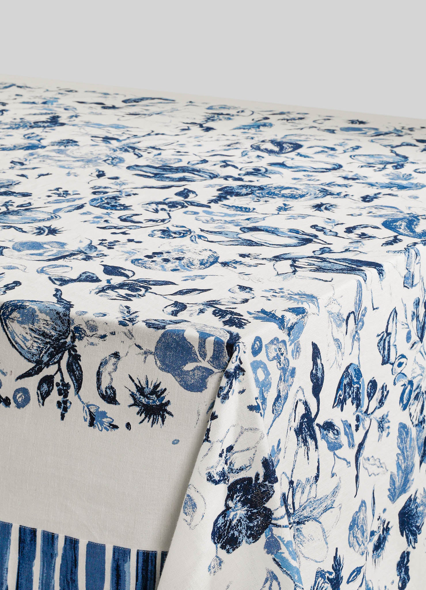 Linen tablecloth with a blue botanical print featuring flowers and leaves on a cream background. The fabric has a soft, natural texture, and the design includes a border with vertical blue stripes.