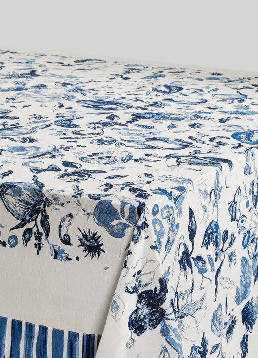Linen tablecloth with a blue botanical print featuring flowers and leaves on a cream background. The fabric has a soft, natural texture, and the design includes a border with vertical blue stripes.