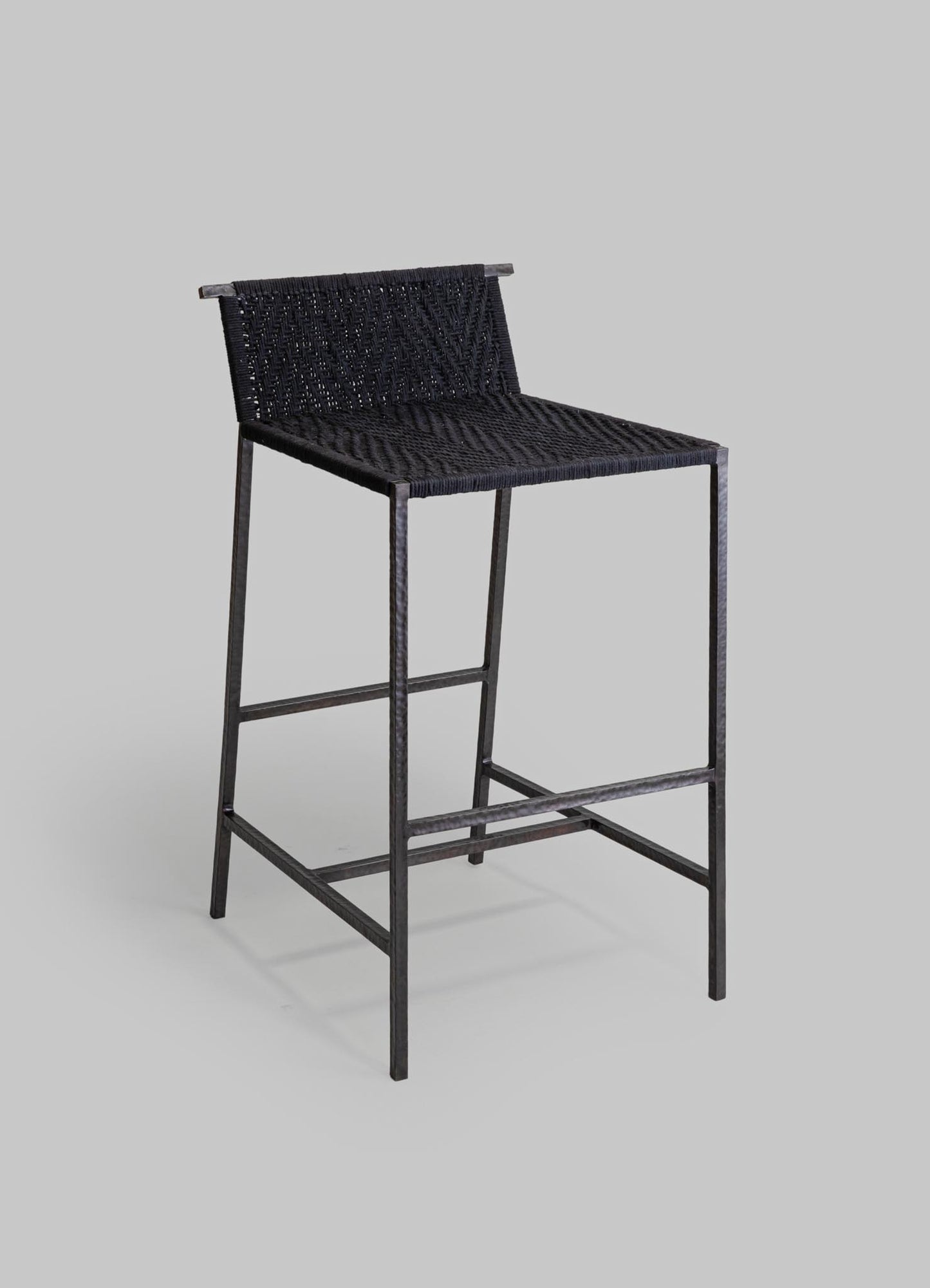 A black woven rattan bar stool with a metal frame, featuring a rectangular seat and a low backrest, set against a plain light gray background.