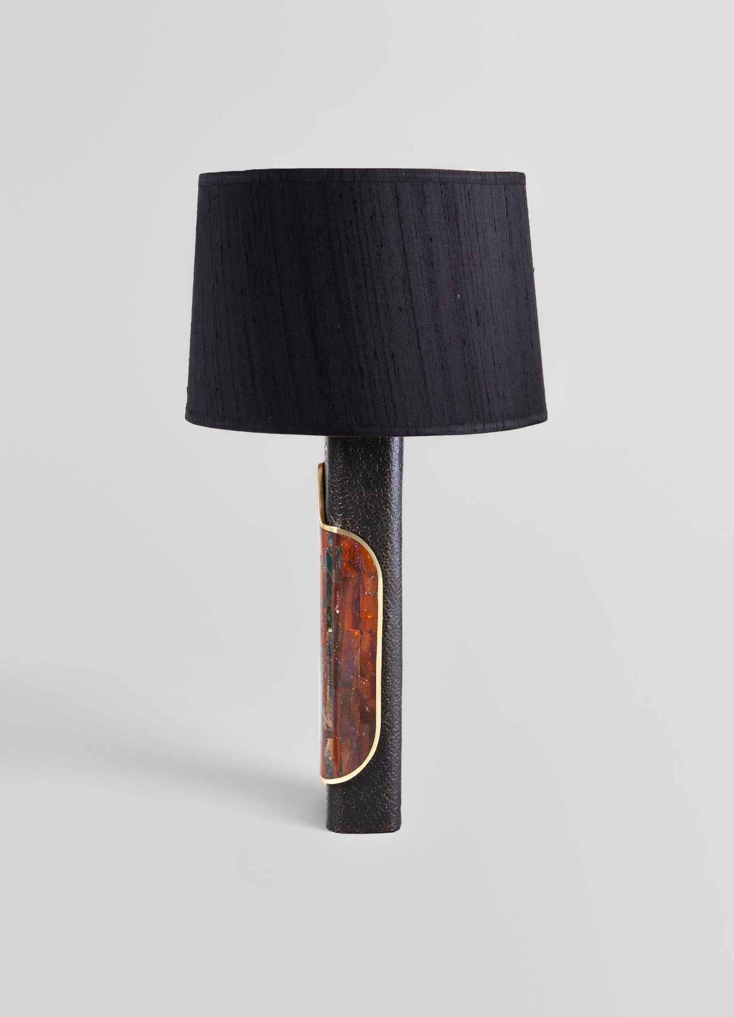 Table lamp with a cylindrical base made of textured dark material, featuring a decorative inlay with orange, green, and red mosaic glass accents edged in gold. The lamp has a black fabric drum shade, creating a contemporary and handcrafted aesthetic.