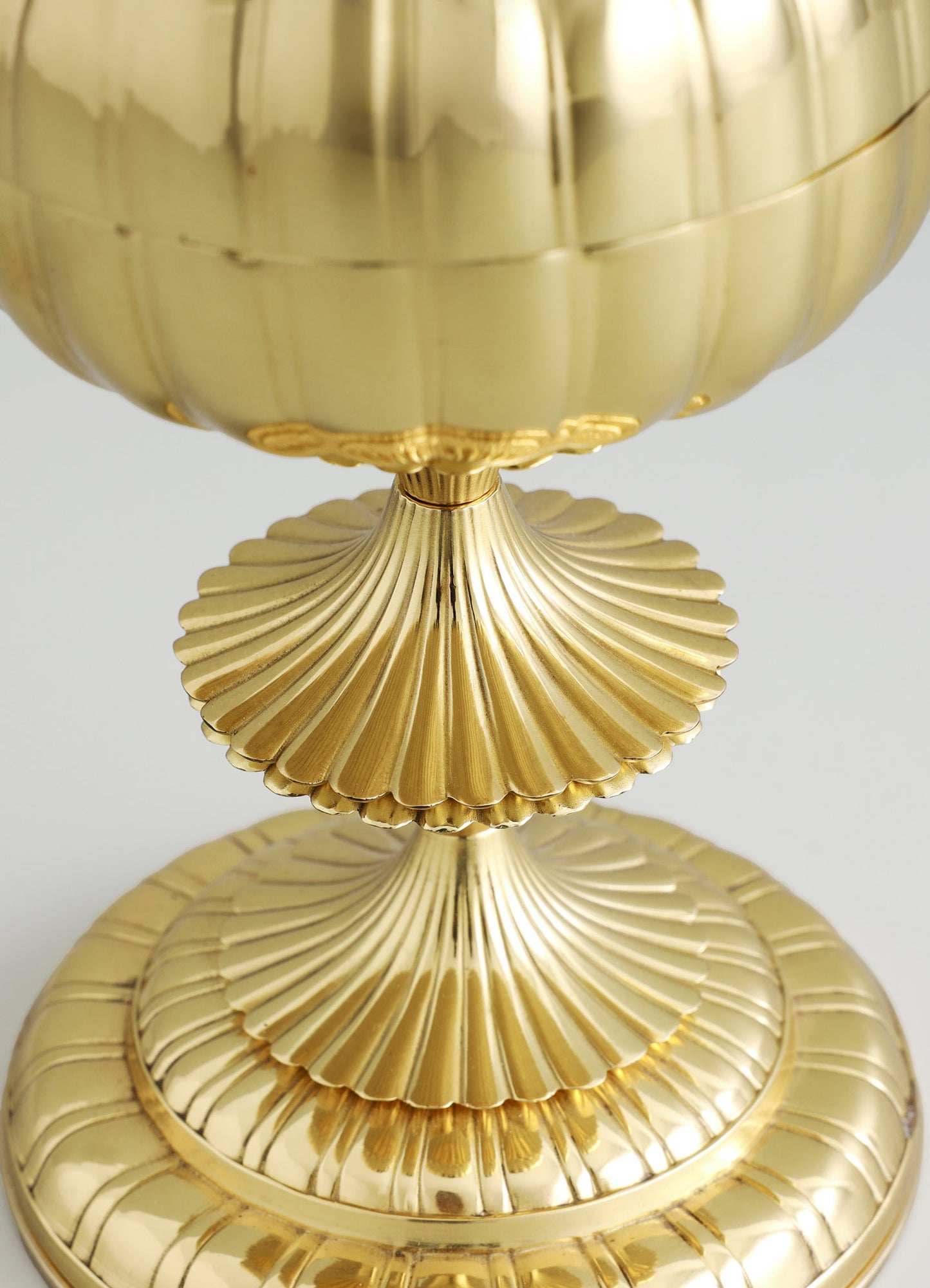 A gold-colored decorative object with a spherical top and a ridged base, featuring a glossy metallic finish and layered, scalloped details on the base. The design has a vintage, ornate style with a polished, reflective surface.