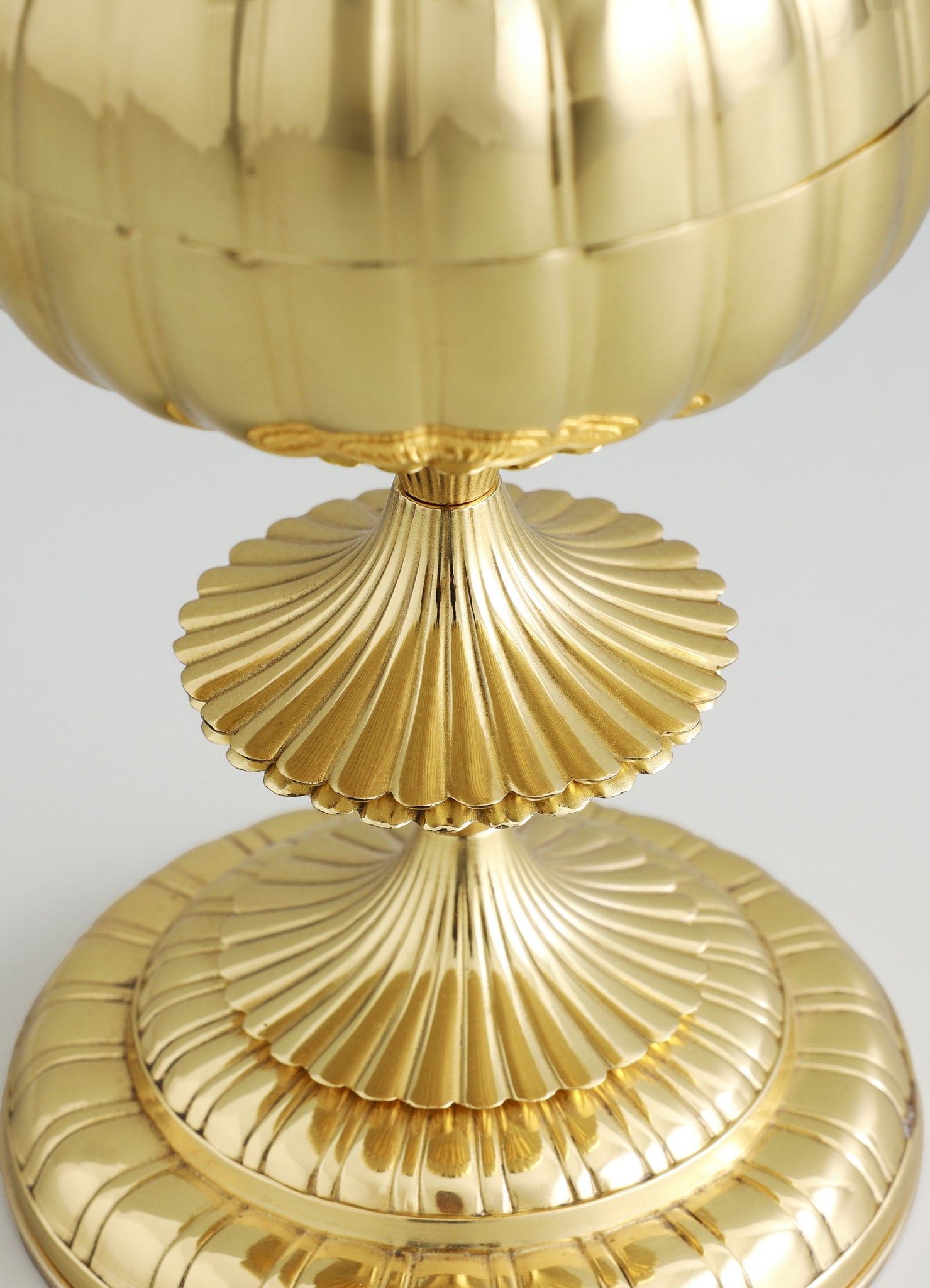 A gold-colored decorative object with a spherical top and a ridged base, featuring a glossy metallic finish and layered, scalloped details on the base. The design has a vintage, ornate style with a polished, reflective surface.