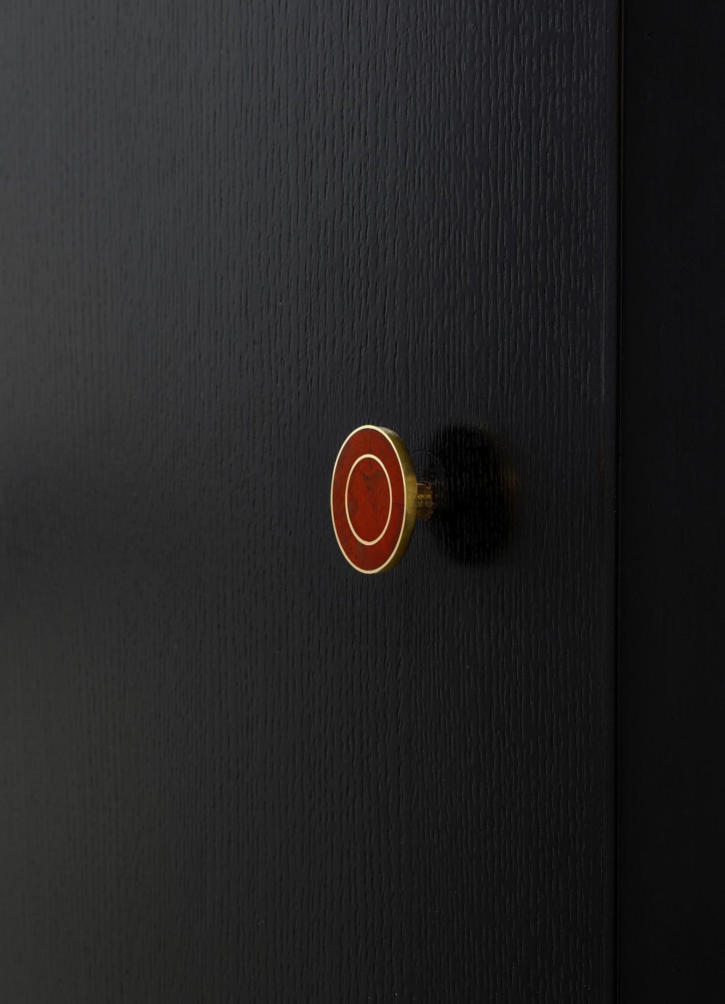 A close-up of a black wooden surface with a small, round brass and red enamel knob featuring white circular accents. The finish of the wood is smooth with a fine grain texture, and the overall style appears modern and minimalist.