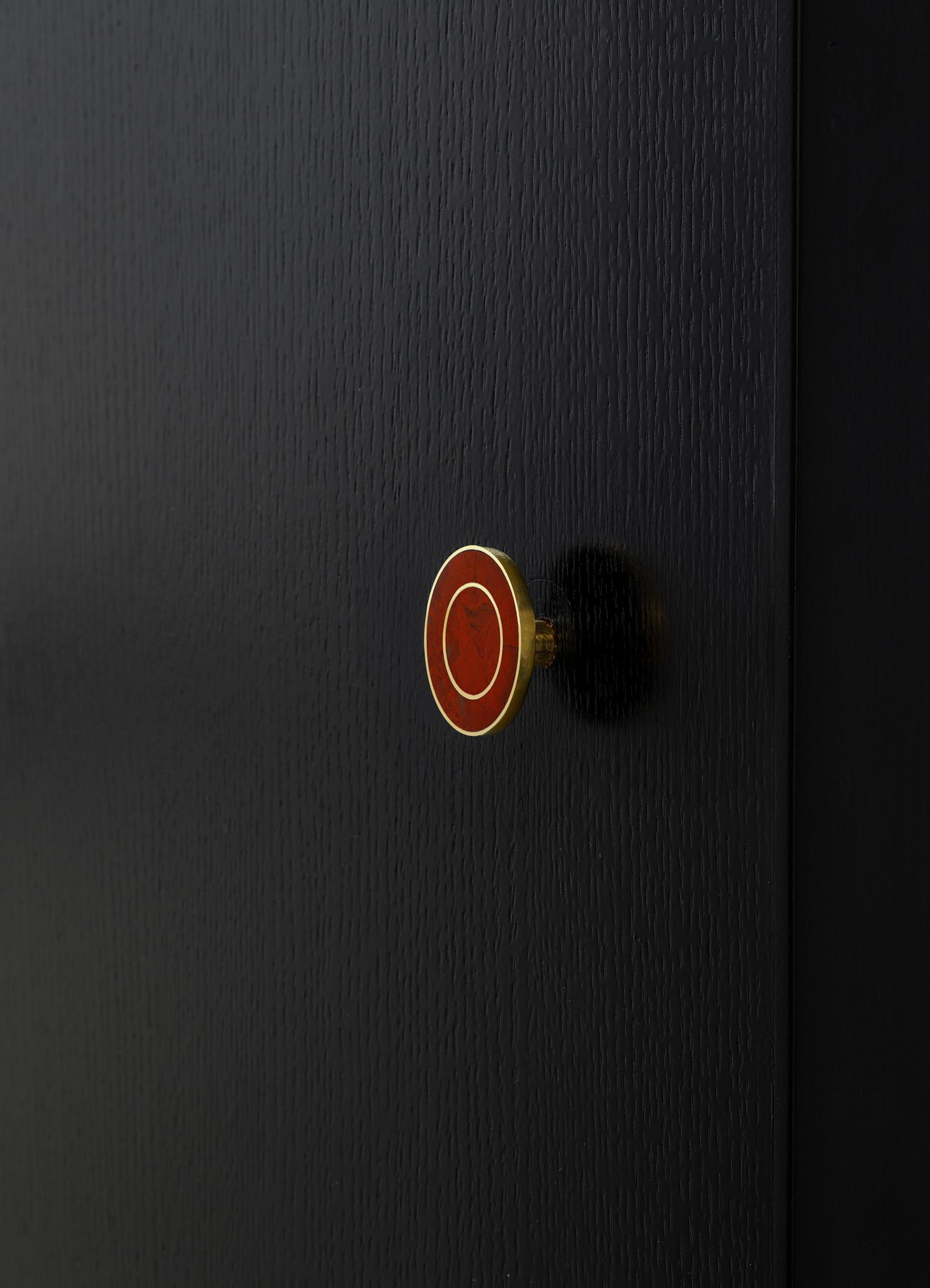 A close-up of a black wooden surface with a small, round brass and red enamel knob featuring white circular accents. The finish of the wood is smooth with a fine grain texture, and the overall style appears modern and minimalist.