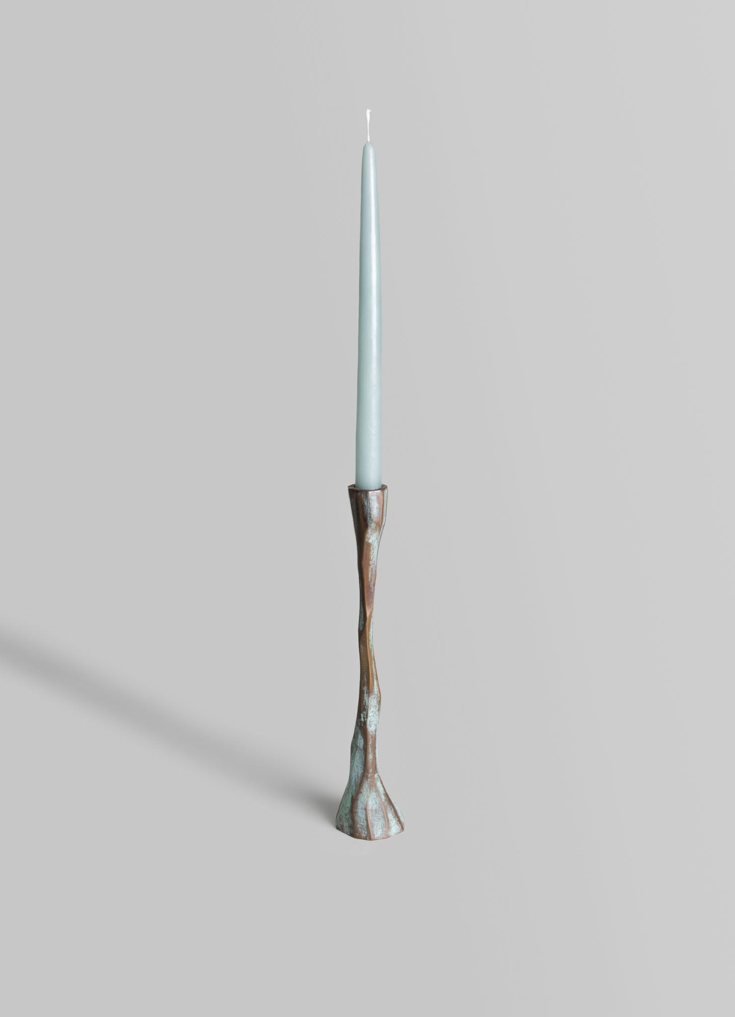 A tall, narrow taper candle holder with a textured, irregular metal base featuring a metallic bronze and patina finish, holding a light blue, slender candle.