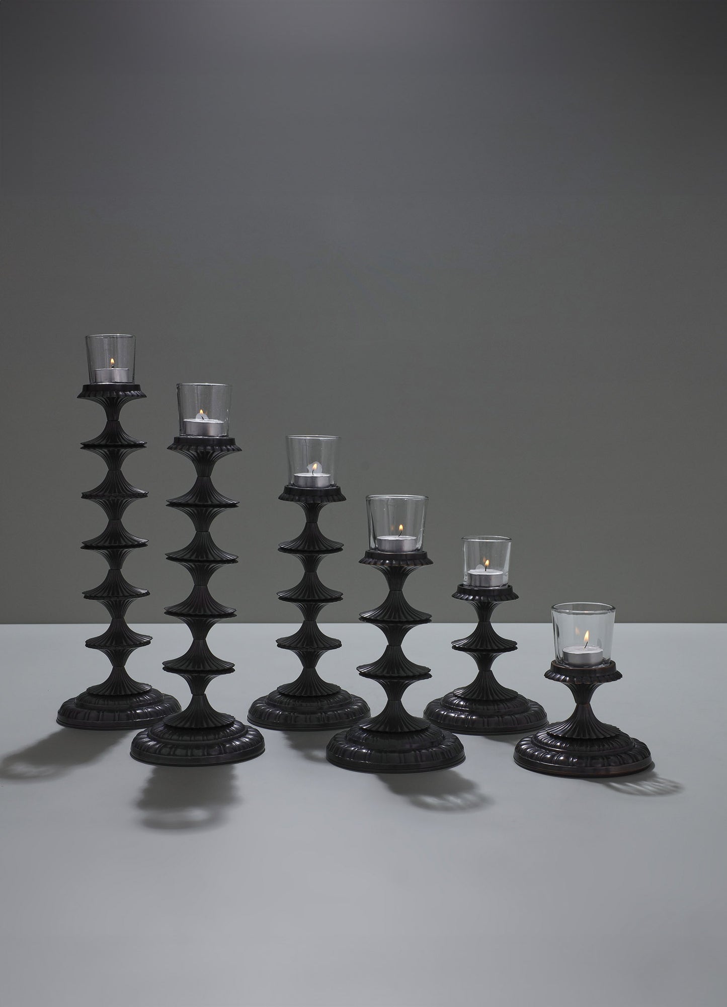 Set of six black candle holders with scalloped, stacked bases and glass votive holders containing lit tealight candles. The candle holders feature a tiered, ornate design and are arranged in descending order on a flat surface with a plain, dark background.