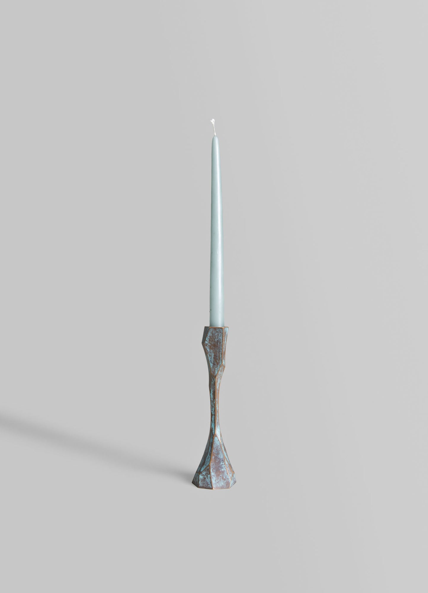 A tall, slender candleholder made of metal with a distressed, oxidized finish in shades of blue and copper. It features a geometric, sculptural design with an asymmetrical base and a tapering form.