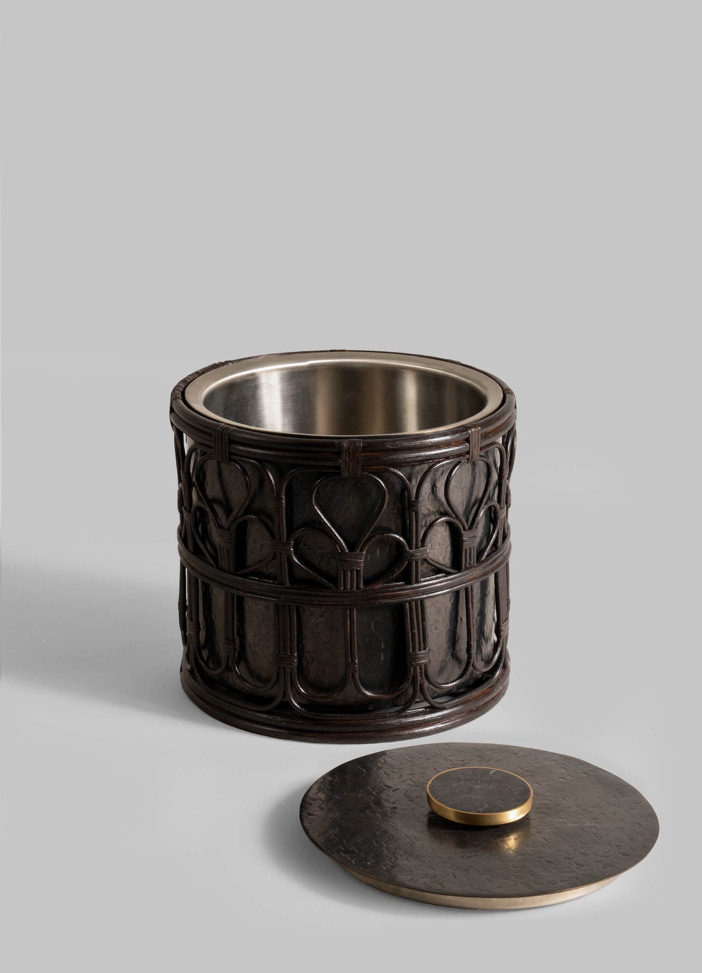 The Manipur Ice Bucket Large by VIYA is a decorative, cylindrical black metal container featuring a cane lattice pattern and metallic lining. Its round lid with a flat handle rests beside it on a plain gray background.