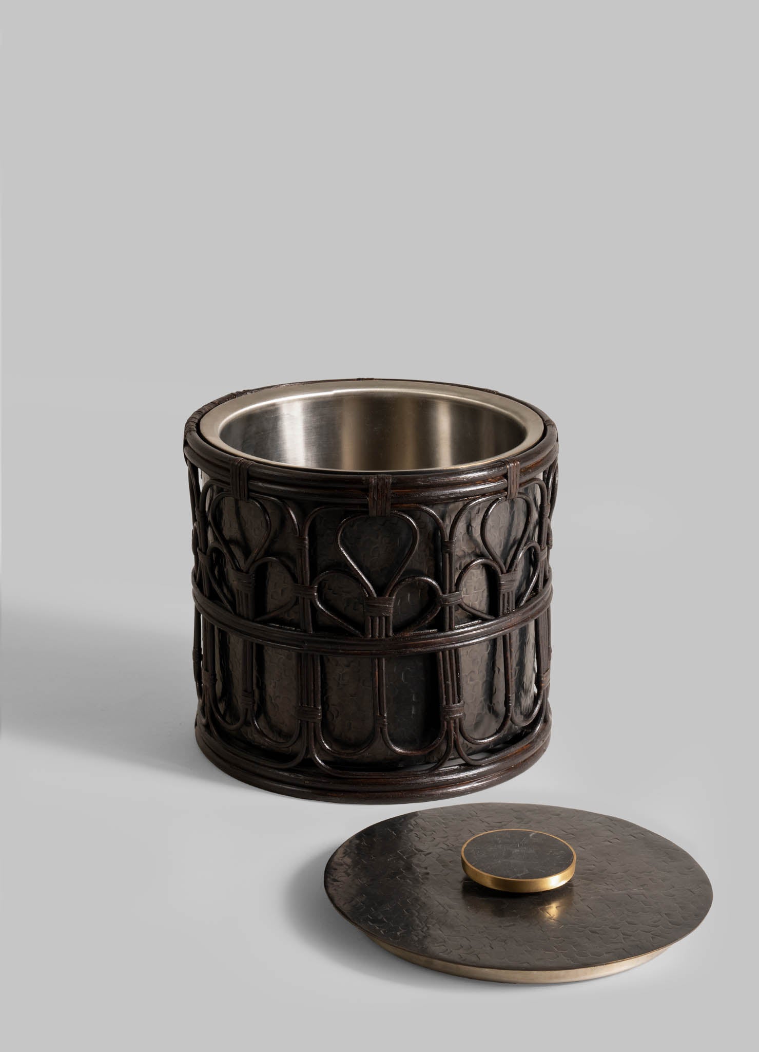 The Manipur Ice Bucket Large by VIYA is a decorative, cylindrical black metal container featuring a cane lattice pattern and metallic lining. Its round lid with a flat handle rests beside it on a plain gray background.