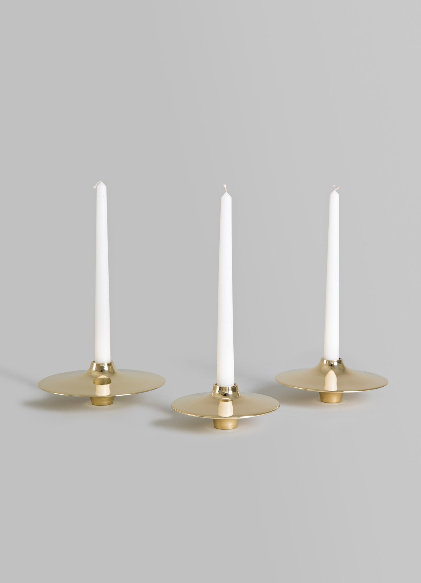 Three brass candle holders with wide, flat circular bases and tall white taper candles, featuring a polished metallic gold finish. The style is minimal and contemporary.
