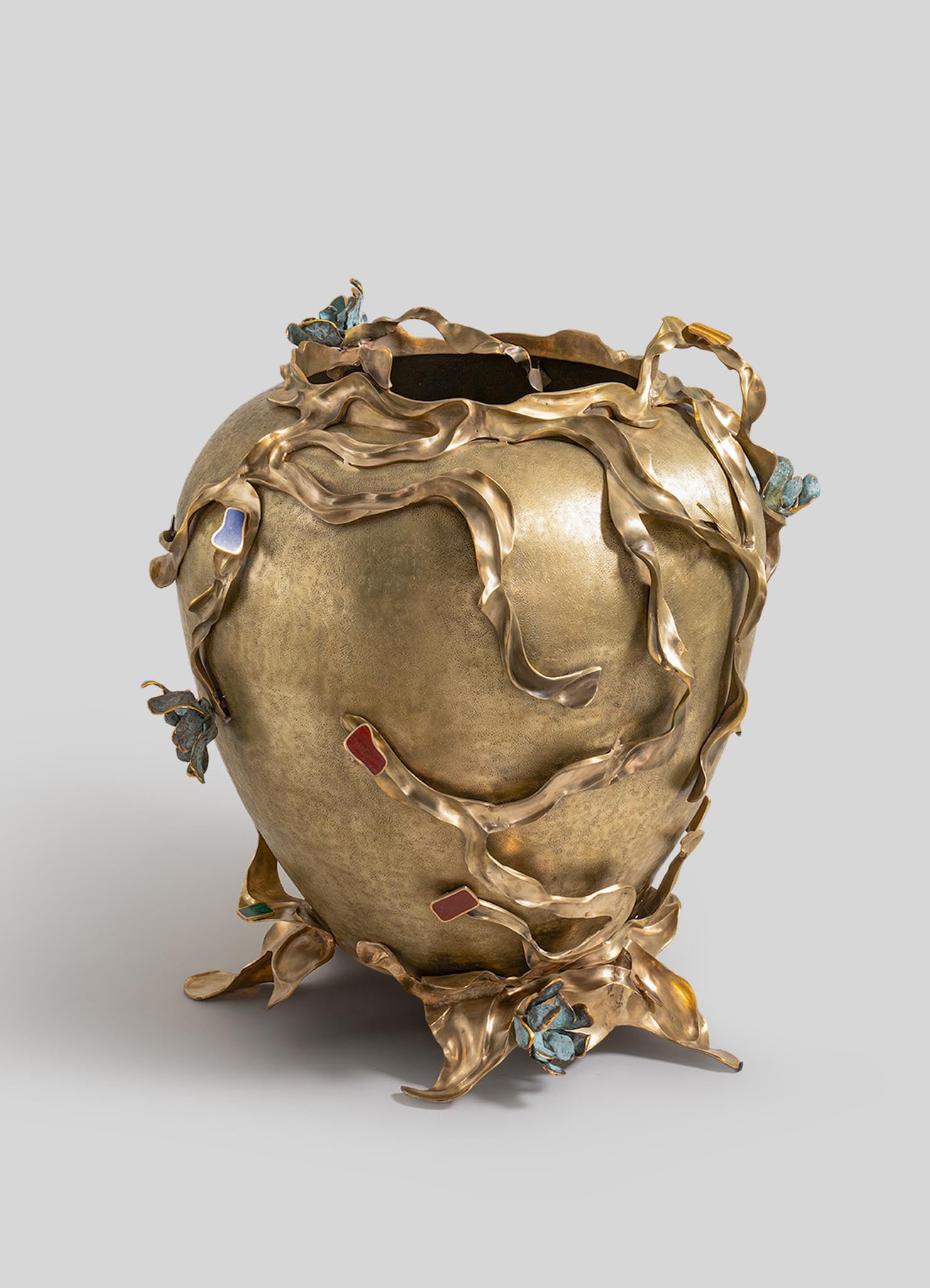 A decorative vase made of metal with a patina finish, featuring organic, flowing leaf-like tendrils wrapping around the body. The tendrils are accented with colorful enamel insets in shades of blue, red, and green, creating a nature-inspired motif.