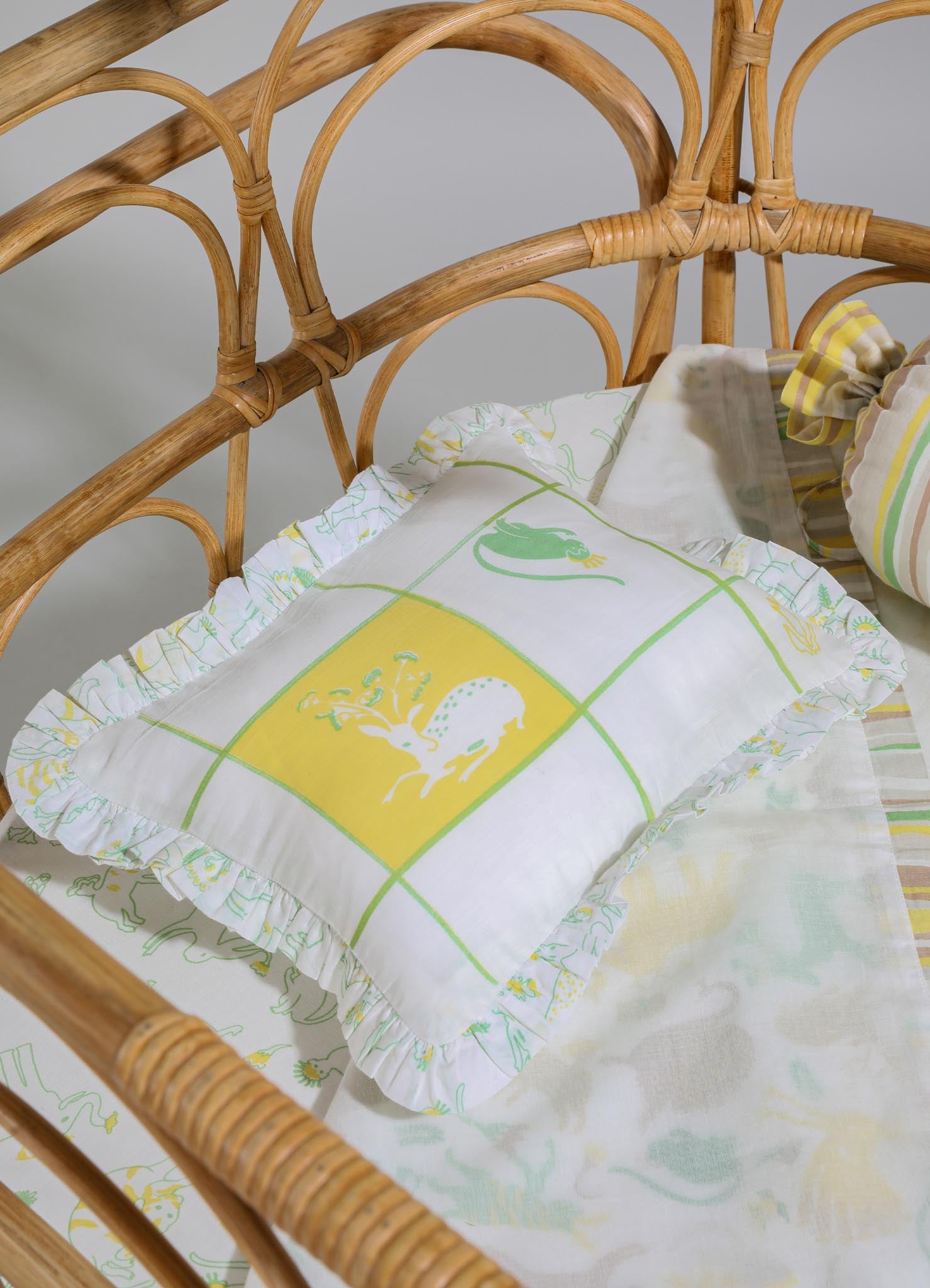 Fabric pillow with a white background, featuring colorful animal and nature-themed motifs in yellow and green, with ruffled edges. Part of a matching bedding set on a rattan bed frame, complemented by striped and patterned linens.