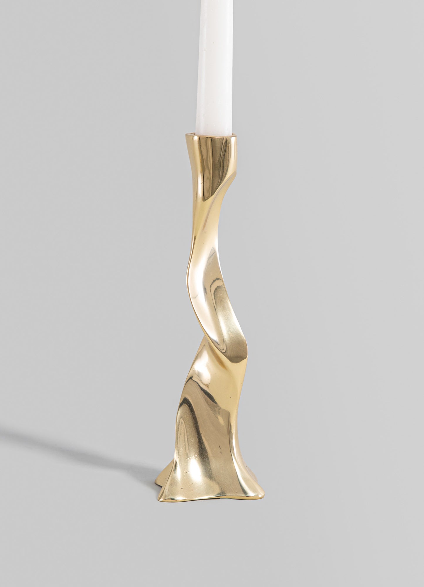 A gold-colored metal candlestick holder with an abstract, sculptural design, holding a white tapered candle. The piece has a smooth, polished surface and a contemporary style.