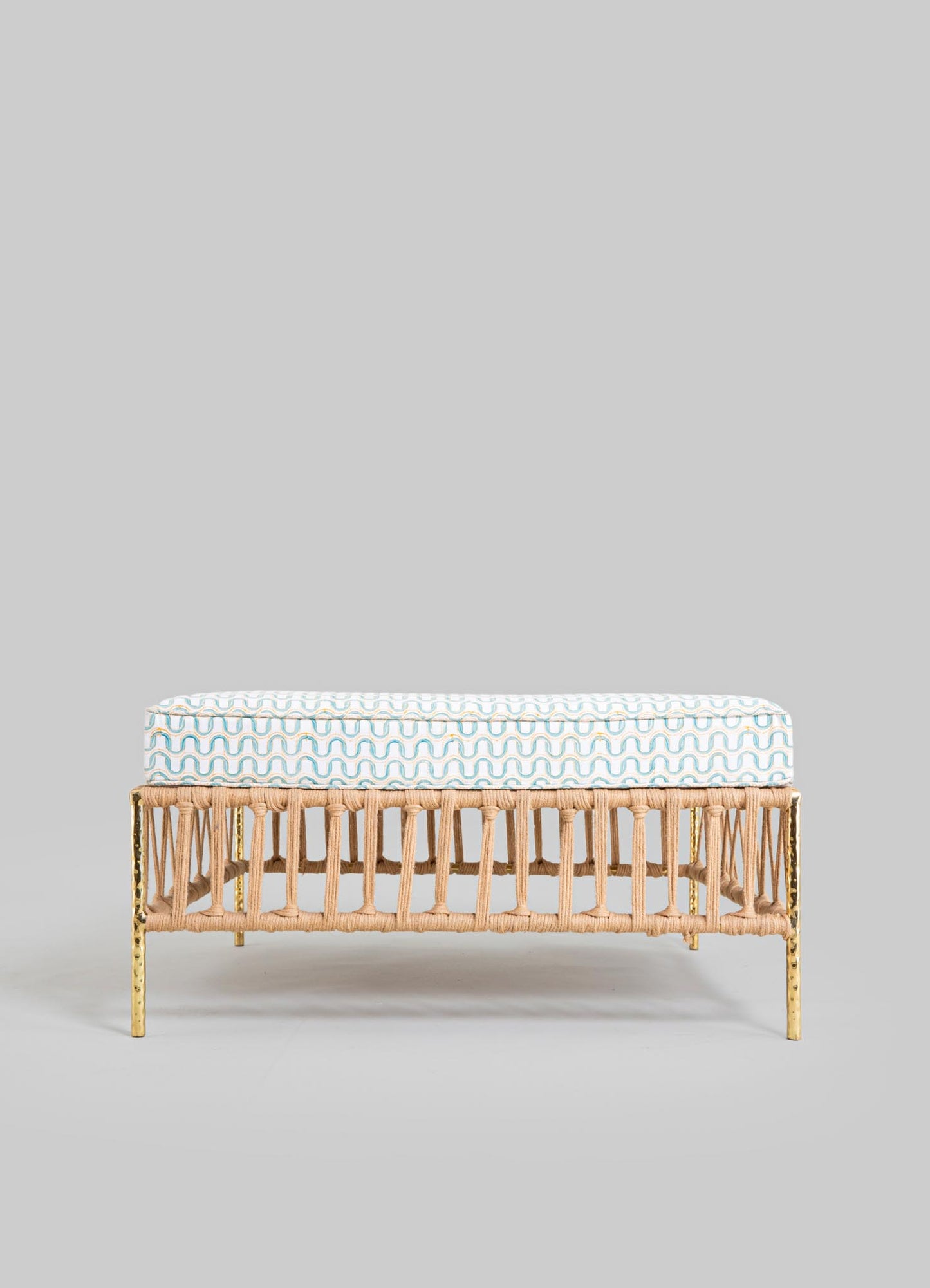 A bench with a rattan frame, featuring a woven design and a metal gold finish on the legs, upholstered with a cushion that has a white background and a wavy blue pattern.