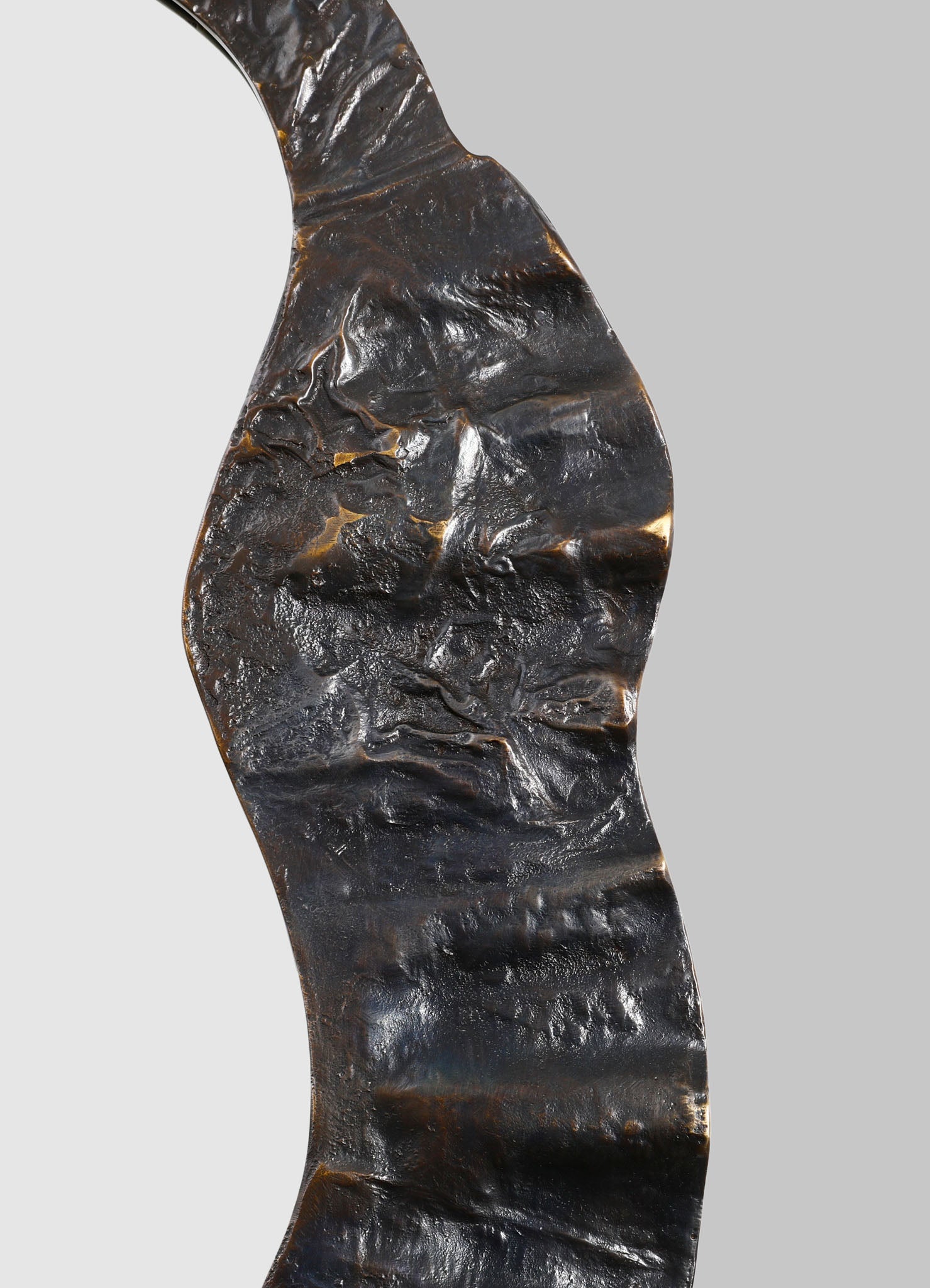 A close-up of a blackened, textured metal sculpture with a wavy, abstract form, featuring a rough, hammered surface and a shiny finish. The sculpture exhibits organic curves and irregularities, emphasizing its handcrafted and contemporary style.