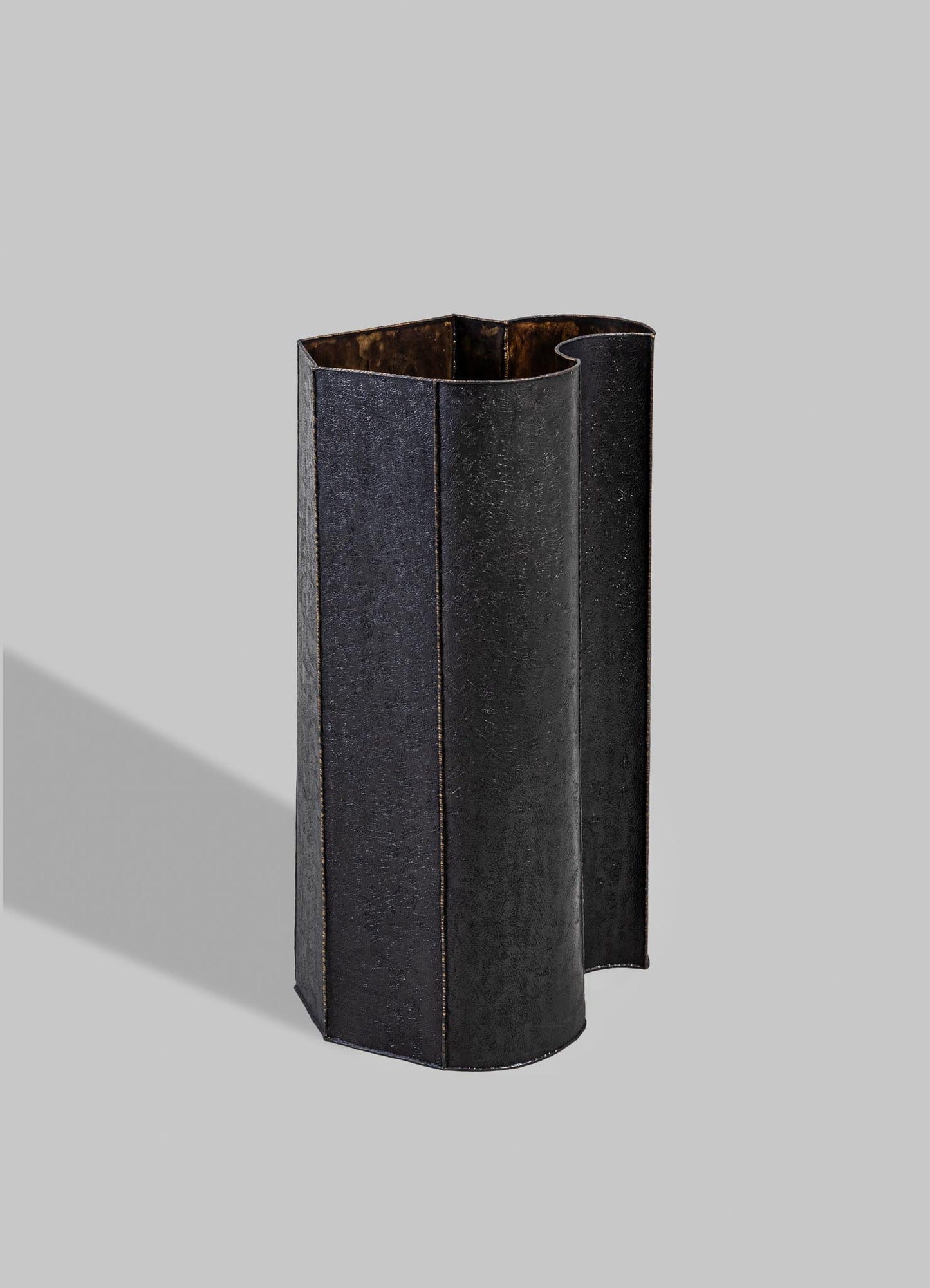 A tall, black metal vase with a textured matte finish and an irregular, curved edge, standing on a neutral gray background.
