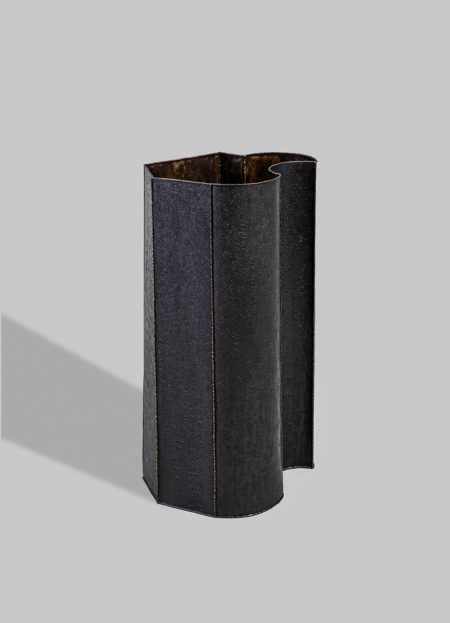 A tall, black metal vase with a textured matte finish and an irregular, curved edge, standing on a neutral gray background.