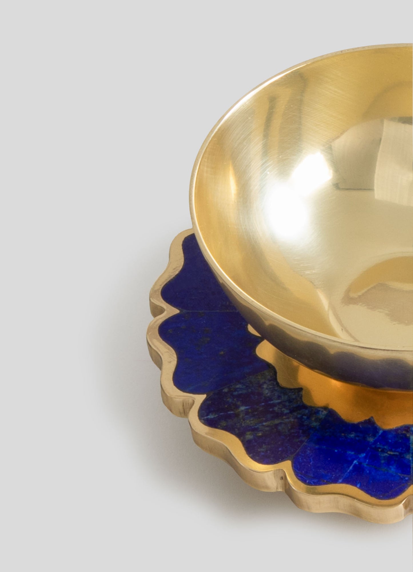 A metallic gold bowl with a smooth, reflective interior placed on a scalloped edge plate featuring a blue and gold finish. The items have a contemporary style with luxurious metallic tones and decorative motifs.