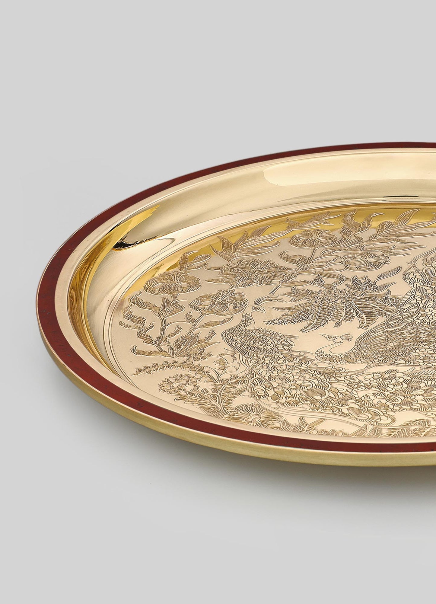 A round decorative tray with a gold-toned metallic surface featuring intricate engraved floral and animal motifs, bordered by a reddish-brown edge.