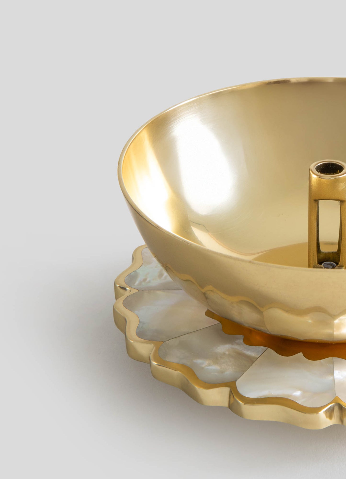 A gold-colored metal bowl with a polished, reflective surface and a rounded shape, placed on a decorative scalloped-edge tray made of similar material with an iridescent, mother-of-pearl finish.