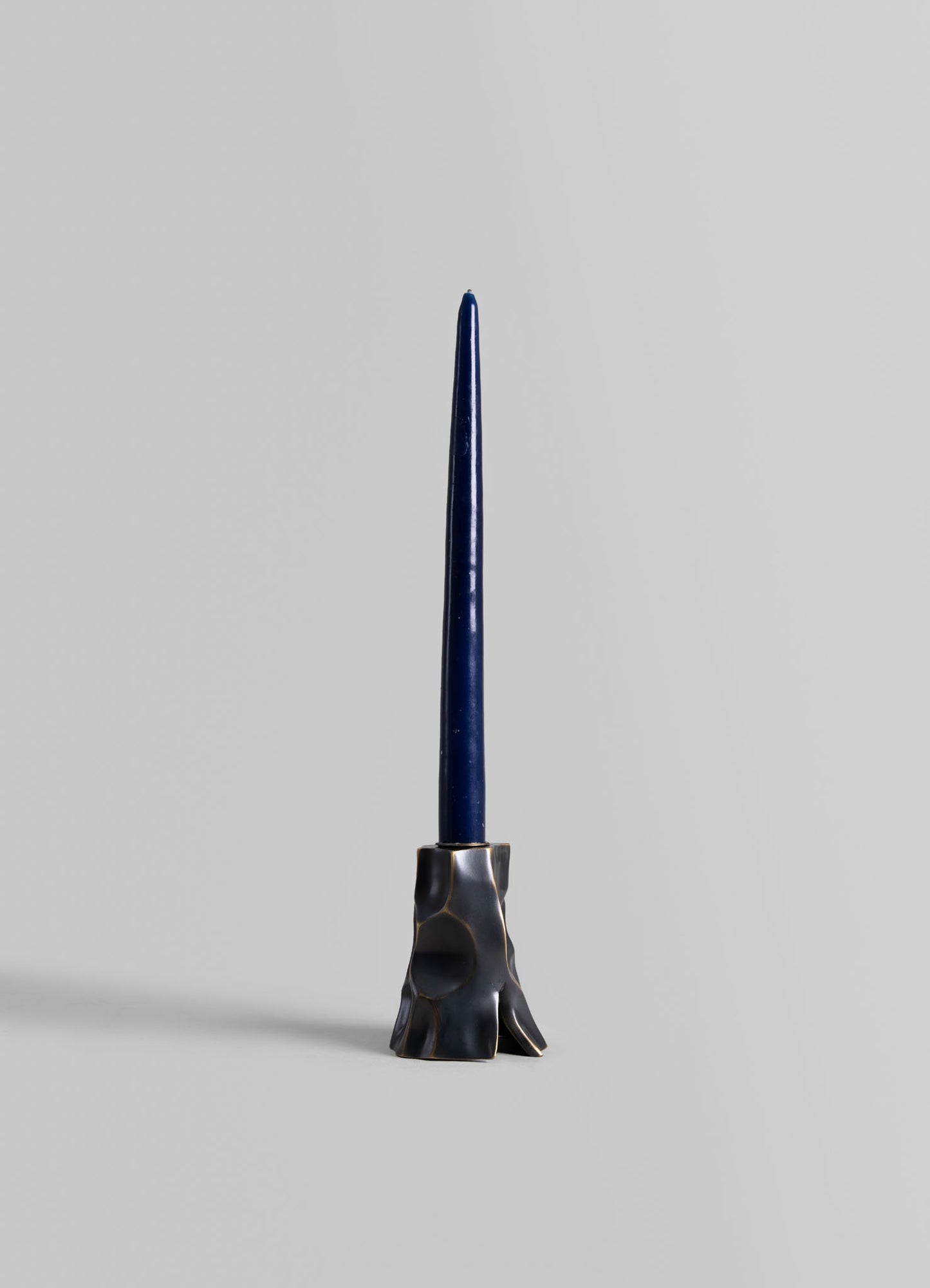 A tall, slender blue candle on a sculptural, metallic holder with an organic, irregular shape and a dark, reflective finish. The candle and holder are set against a plain, neutral background.