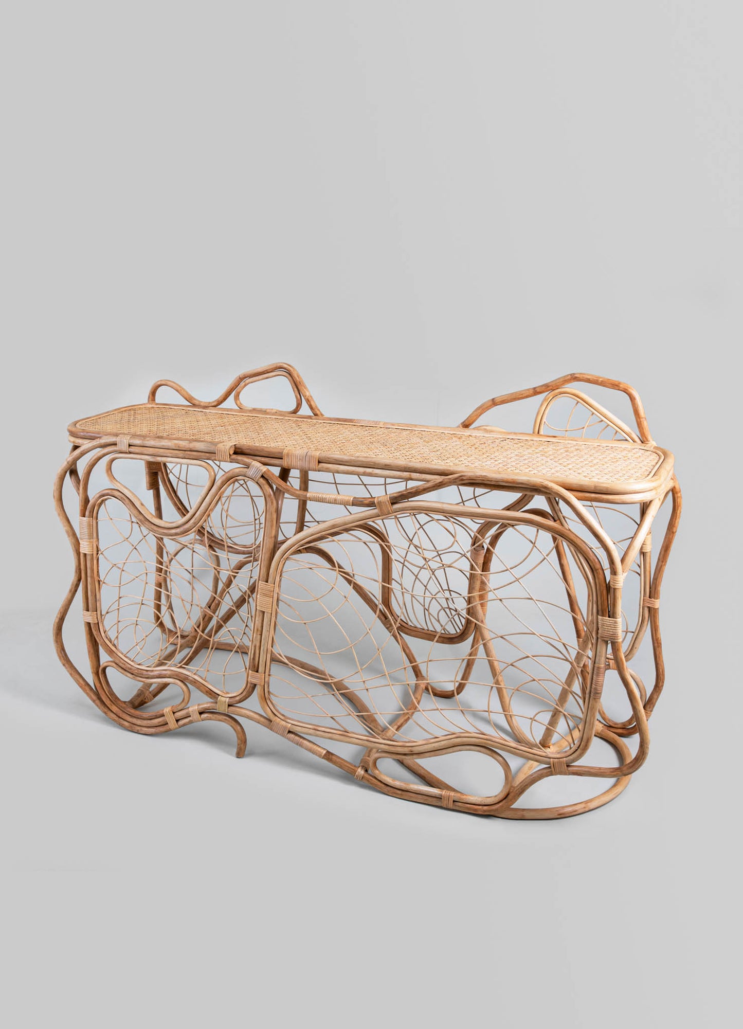 Woven rattan side table with a curved, openwork design featuring intricate loops and circular motifs, finished in a natural, light wood tone. The table has an elongated rectangular shape with a flat top surface and a handcrafted, rustic style.