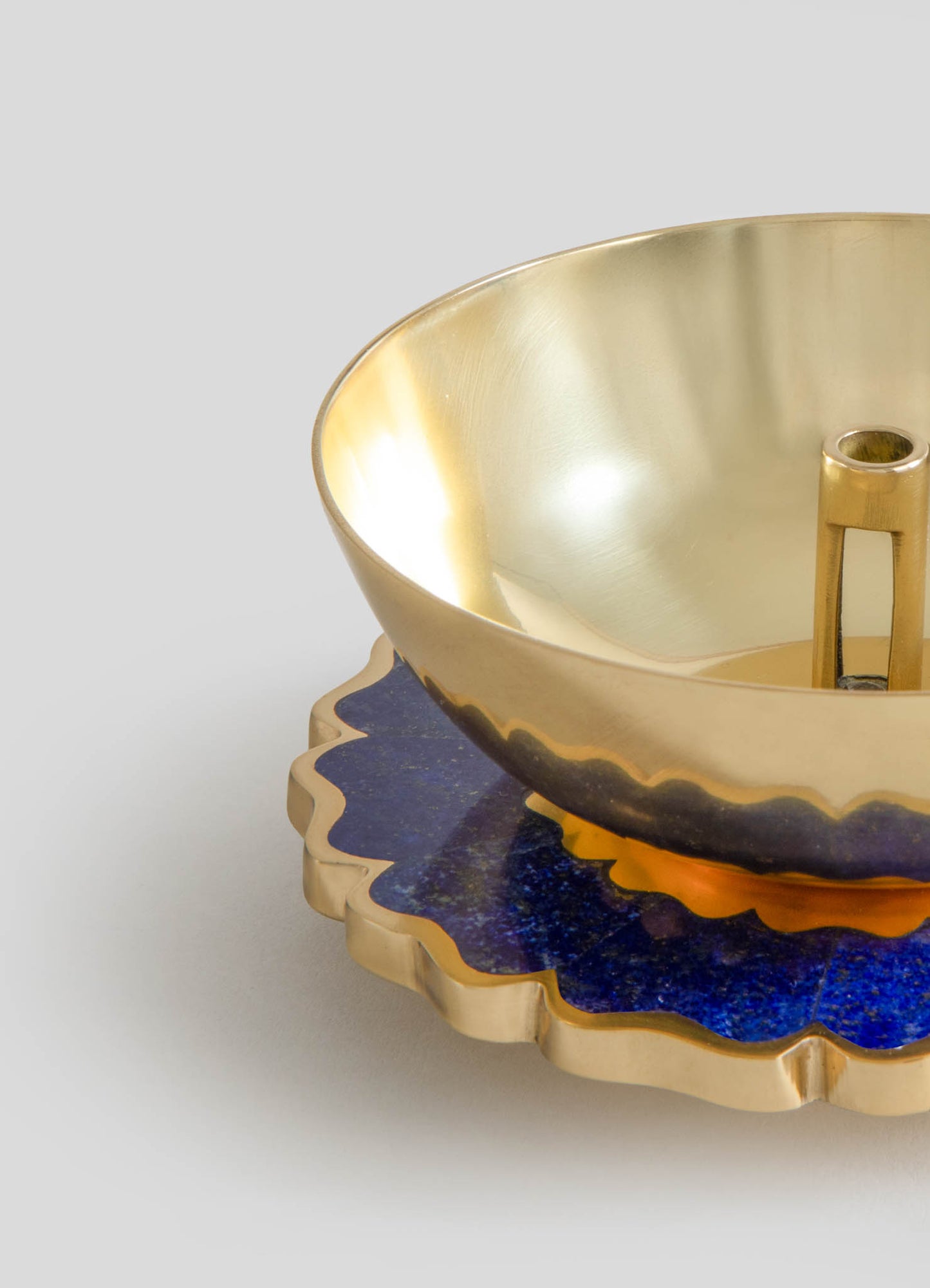 A decorative gold bowl with a smooth, reflective interior and a flared rim sits on a patterned tray with scalloped edges, featuring a deep blue and gold finish. The tray appears to be made of metal with a textured surface that enhances its ornate design.