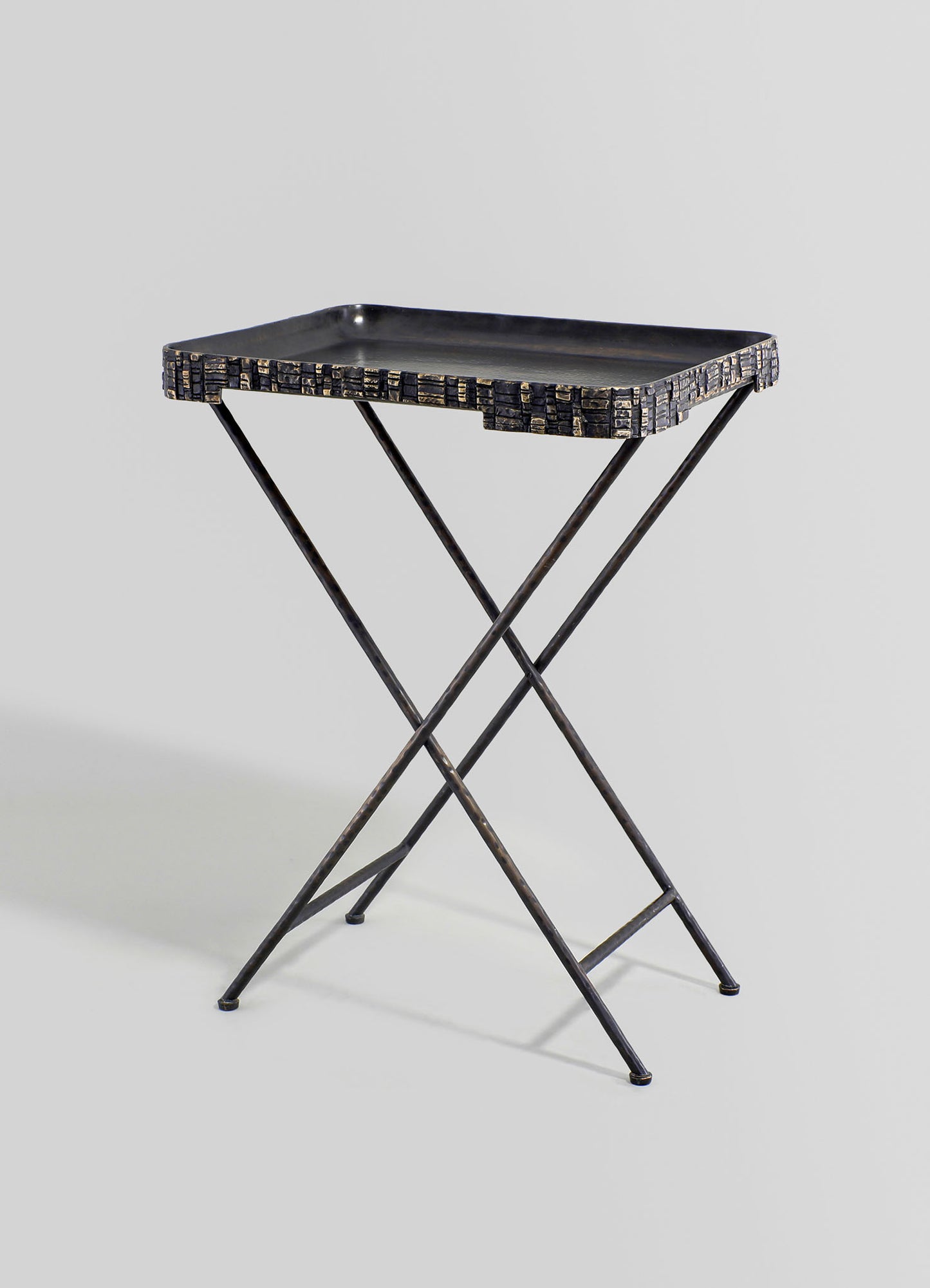 Metal serving tray with a black finish, with textured mosaic-like edges, on a metal stand with crossed legs in a minimalist style.
