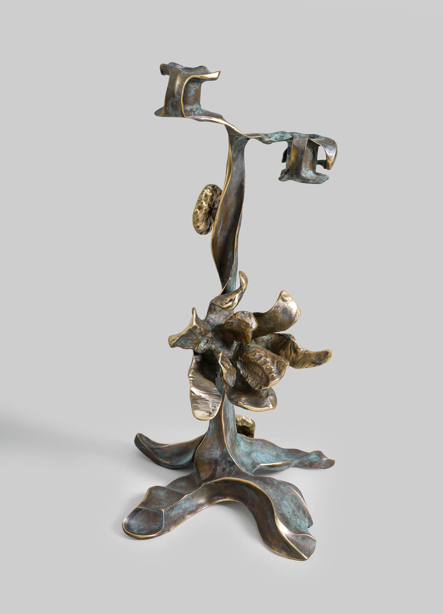 A sculptural decorative object made of metal with a bronze finish, featuring abstract, organic shapes and layered textures, set against a plain gray background.