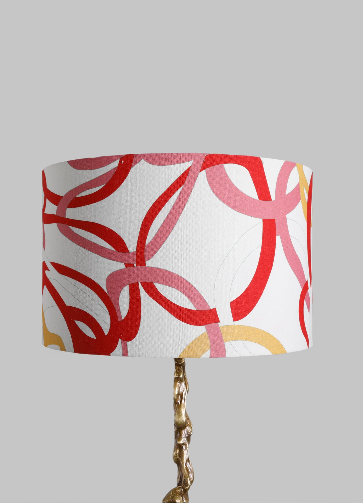 Table lamp with a fabric drum shade featuring a multicolored abstract pattern of intersecting loops in red, pink, and yellow tones. The base appears to be metallic with a textured, irregular gold finish, creating a contemporary and artistic style.