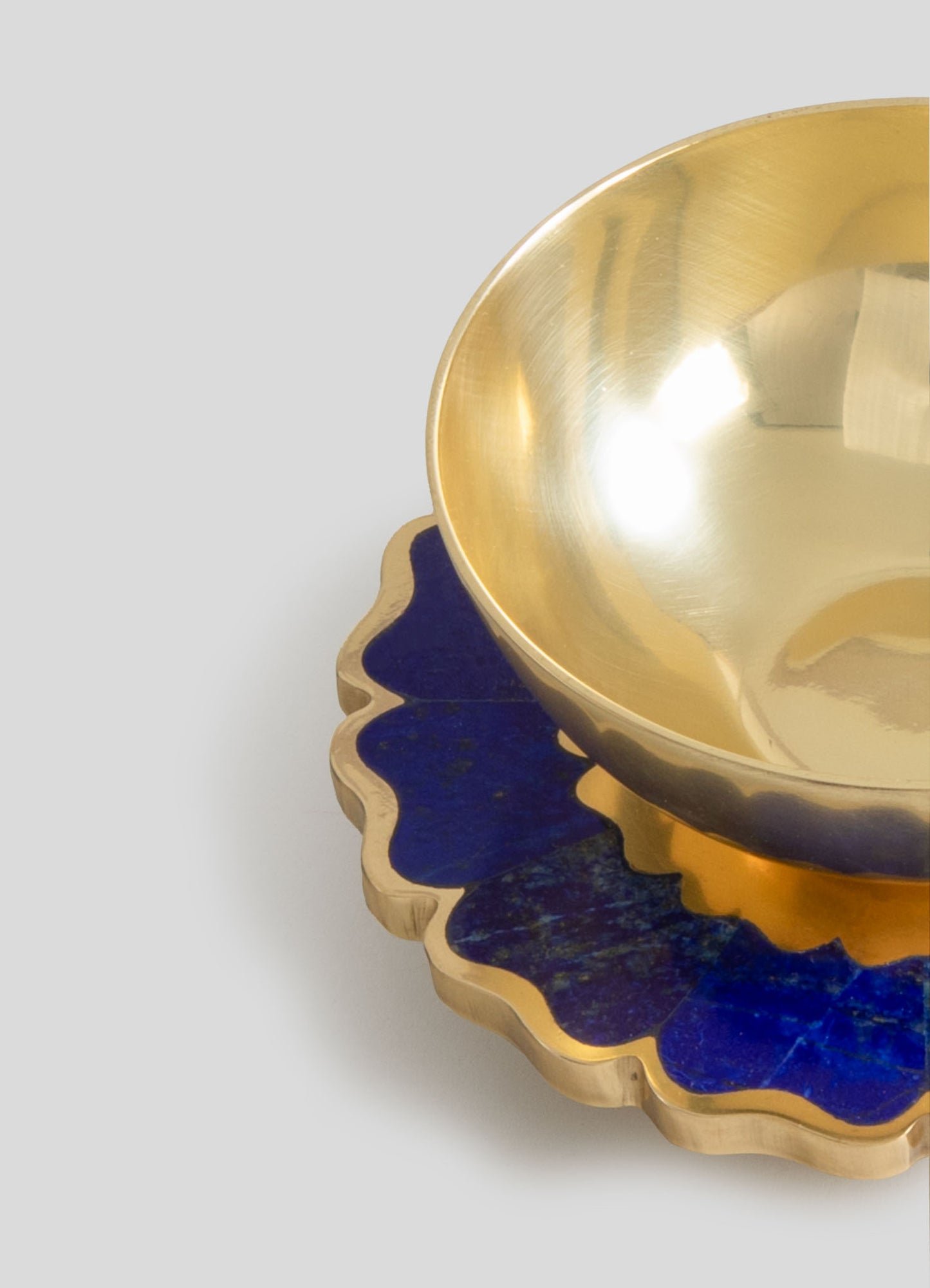 A gold-toned metal bowl with a smooth, reflective interior surface sits atop a decorative tray with a wavy, scalloped edge and a deep blue finish. Both items feature a luxurious, contemporary style with sleek lines and contrasting textures.