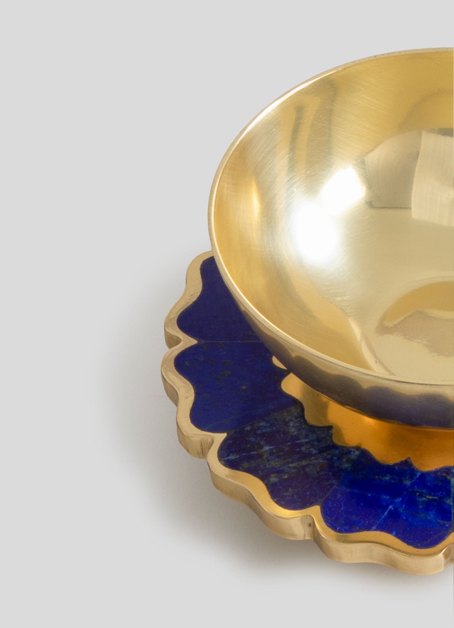 A gold-toned metal bowl with a smooth, reflective interior surface sits atop a decorative tray with a wavy, scalloped edge and a deep blue finish. Both items feature a luxurious, contemporary style with sleek lines and contrasting textures.