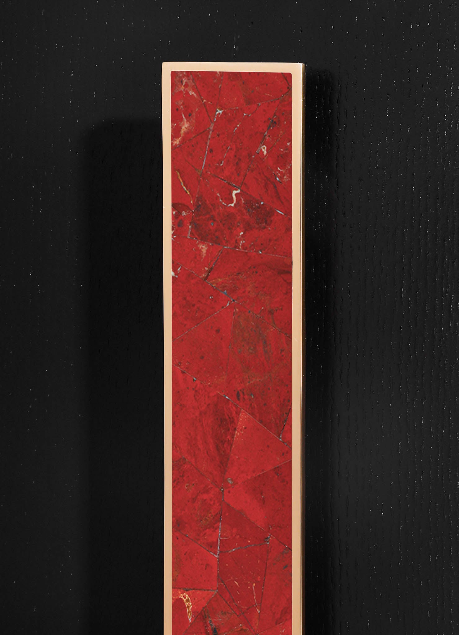A tall, rectangular marble sculpture with a red and black veined pattern, framed by a thin beige border, set against a black textured background.