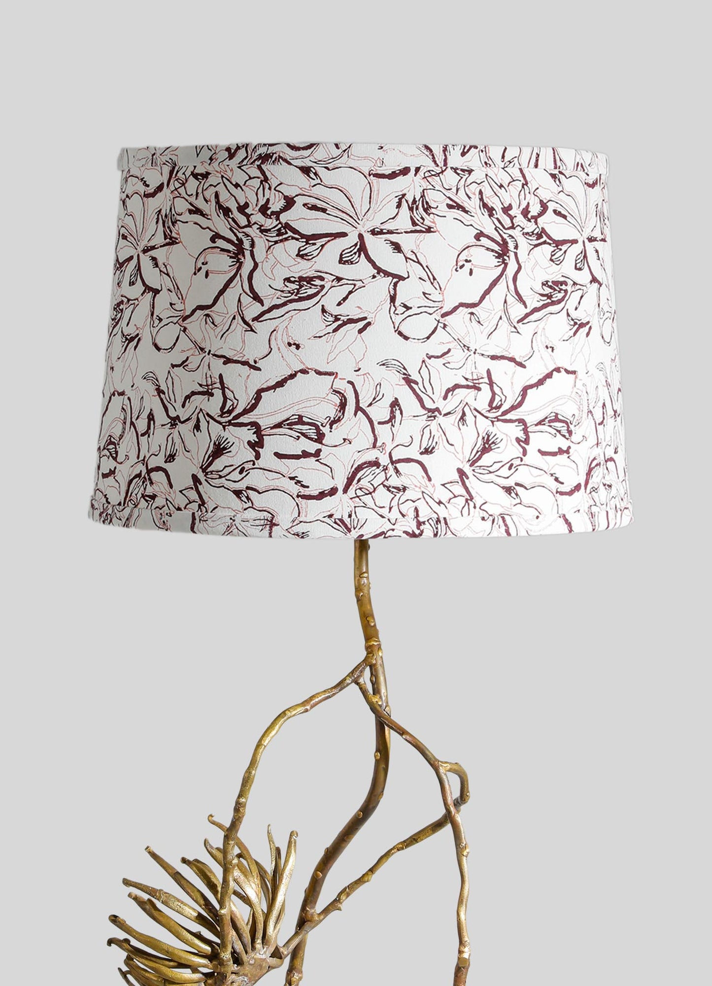 Decorative table lamp with a drum-shaped shade featuring an abstract floral pattern in shades of burgundy and cream, supported by a sculptural base made of twisted, natural wood branches with a gold finish.