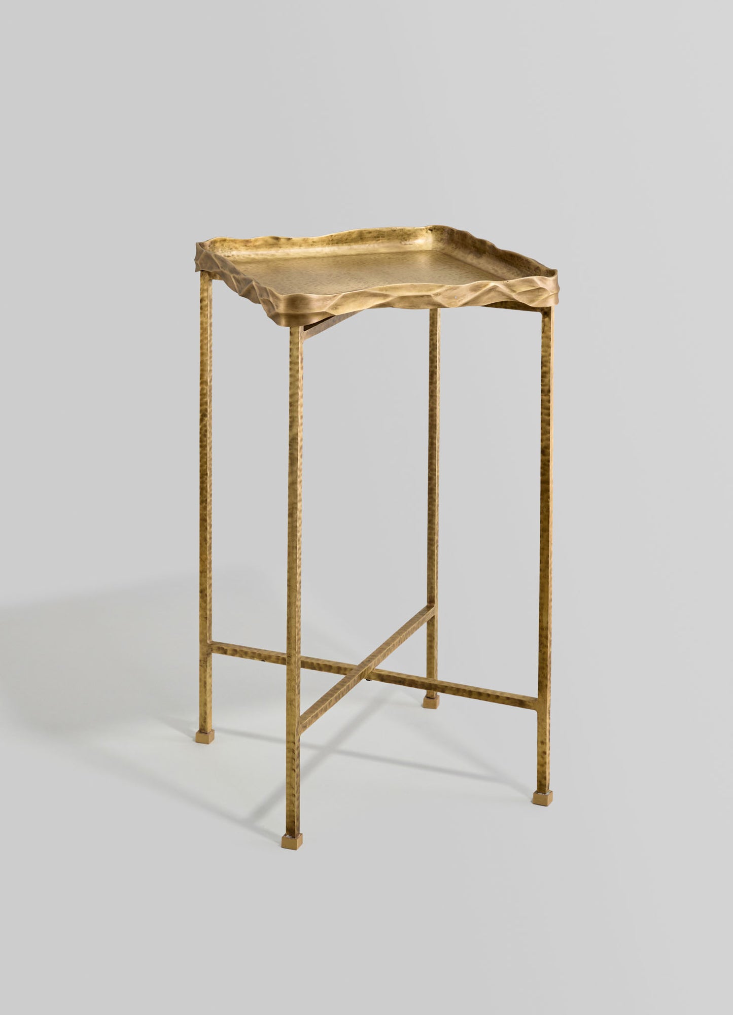 A brass side table with hammered texture and a rectangular top with a wavy, irregular edge, supported by slender, hammered brass legs on a light gray background.