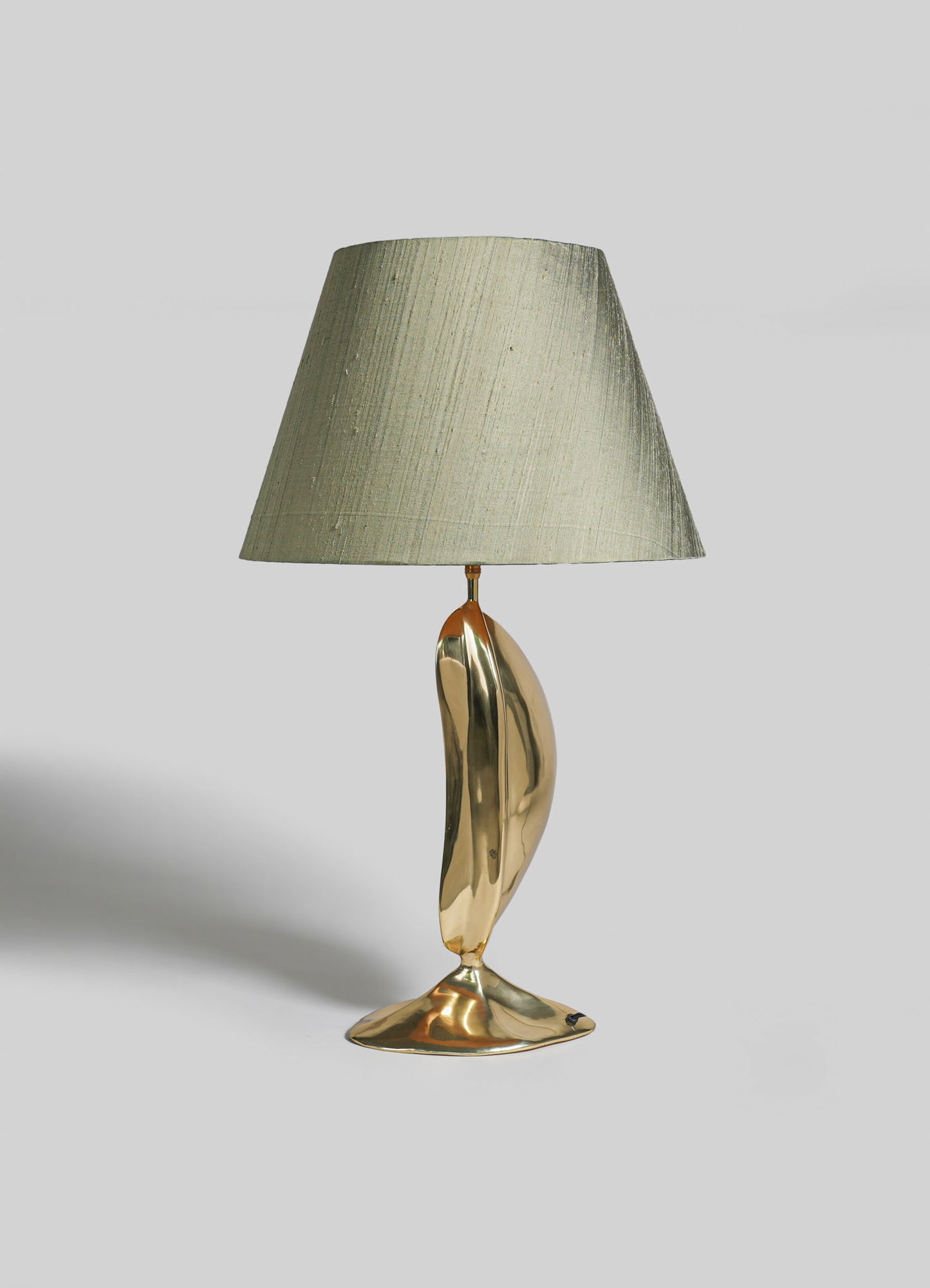 A table lamp with a sculptural brass base and an oval shape, topped with a neutral-colored fabric lampshade. The design exhibits contemporary and artistic style with polished metallic finish and organic form.