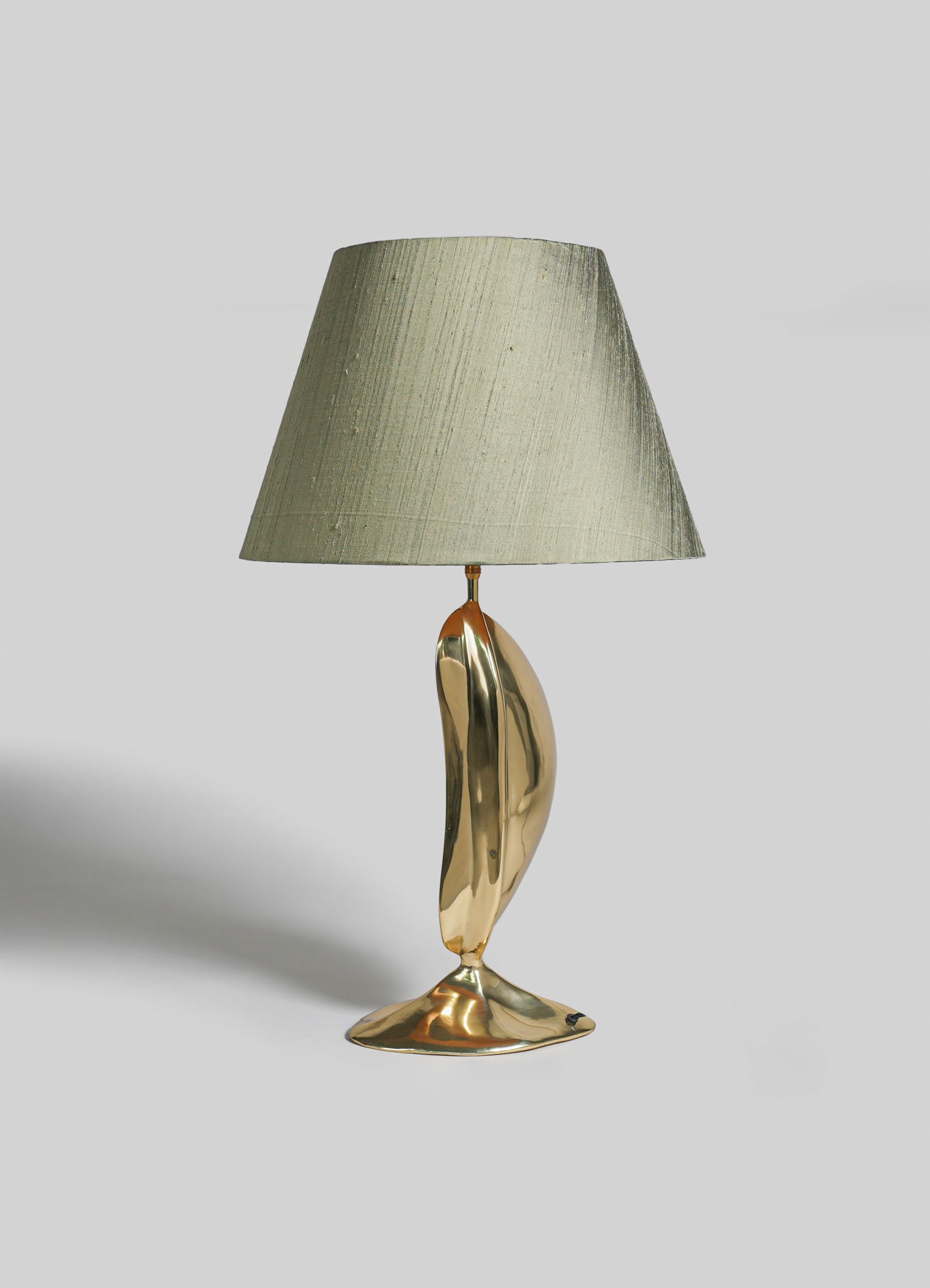 A table lamp with a sculptural brass base and an oval shape, topped with a neutral-colored fabric lampshade. The design exhibits contemporary and artistic style with polished metallic finish and organic form.