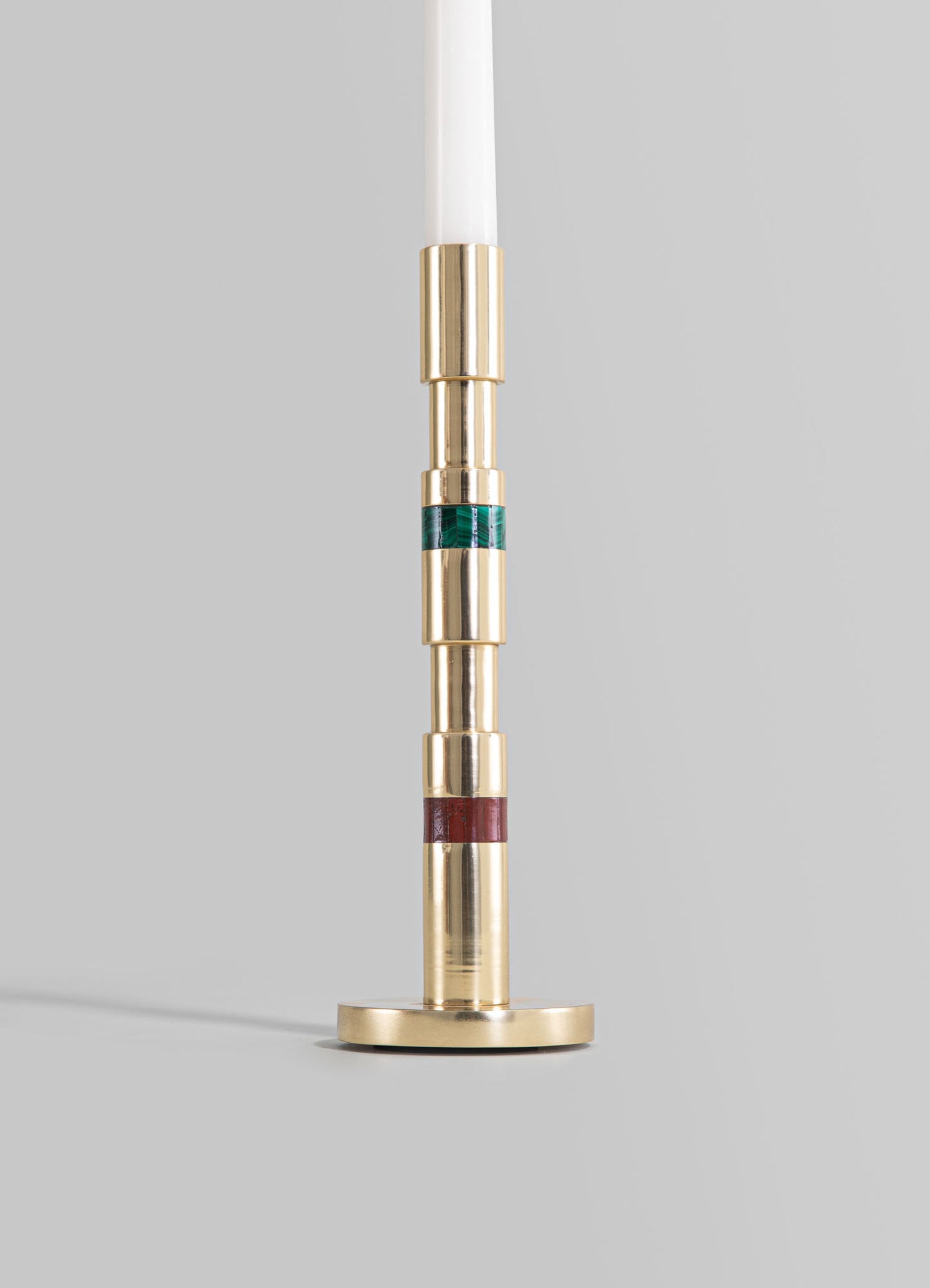 A brass candle holder with a cylindrical design featuring colorful enamel bands in green, red, and uncolored sections, mounted on a flat circular base with a white taper candle inserted.